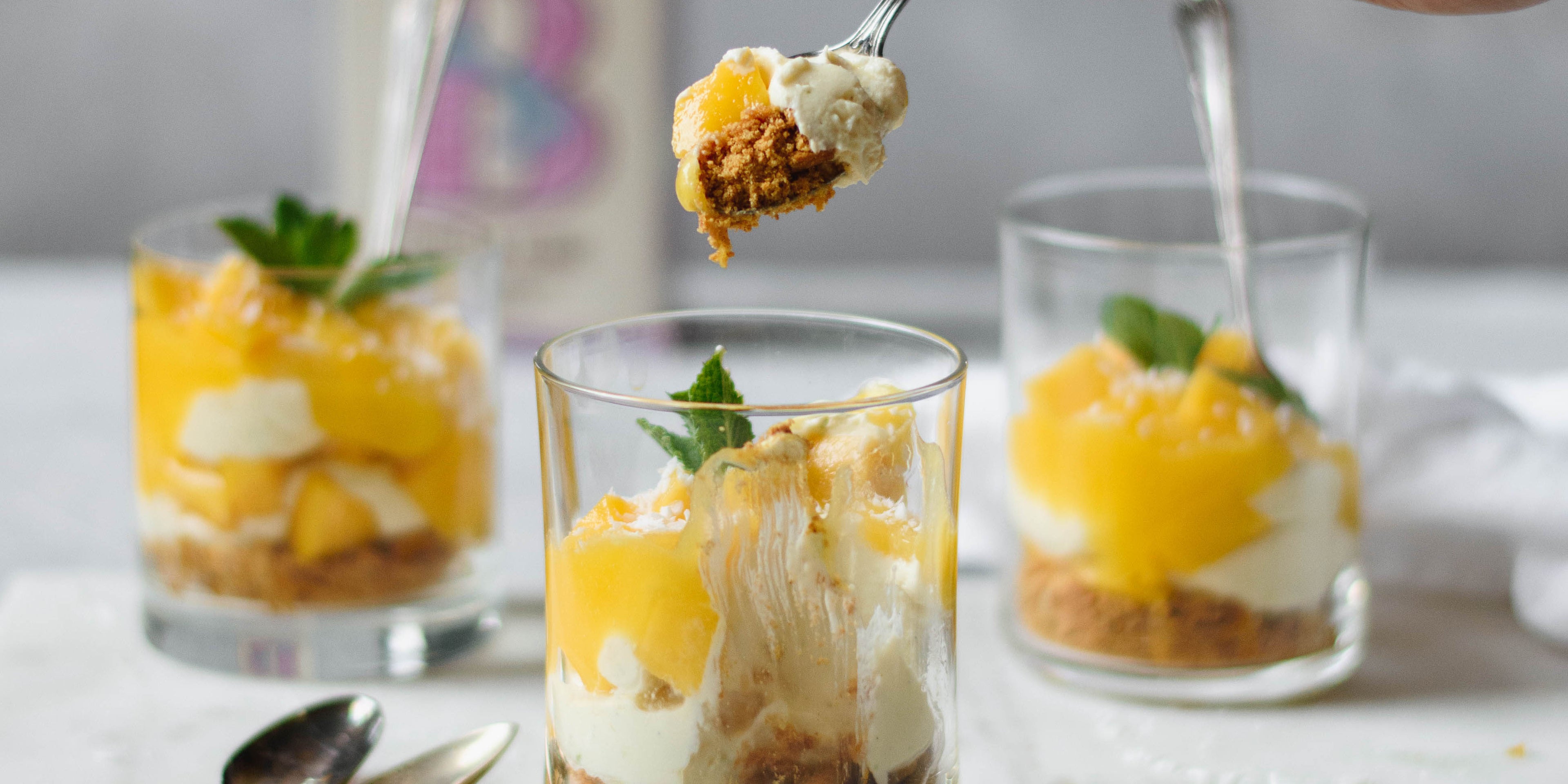 3 small glasses of mango syllabub with sprig of mint on top and two spoons. Spoonful of dessert scooped out