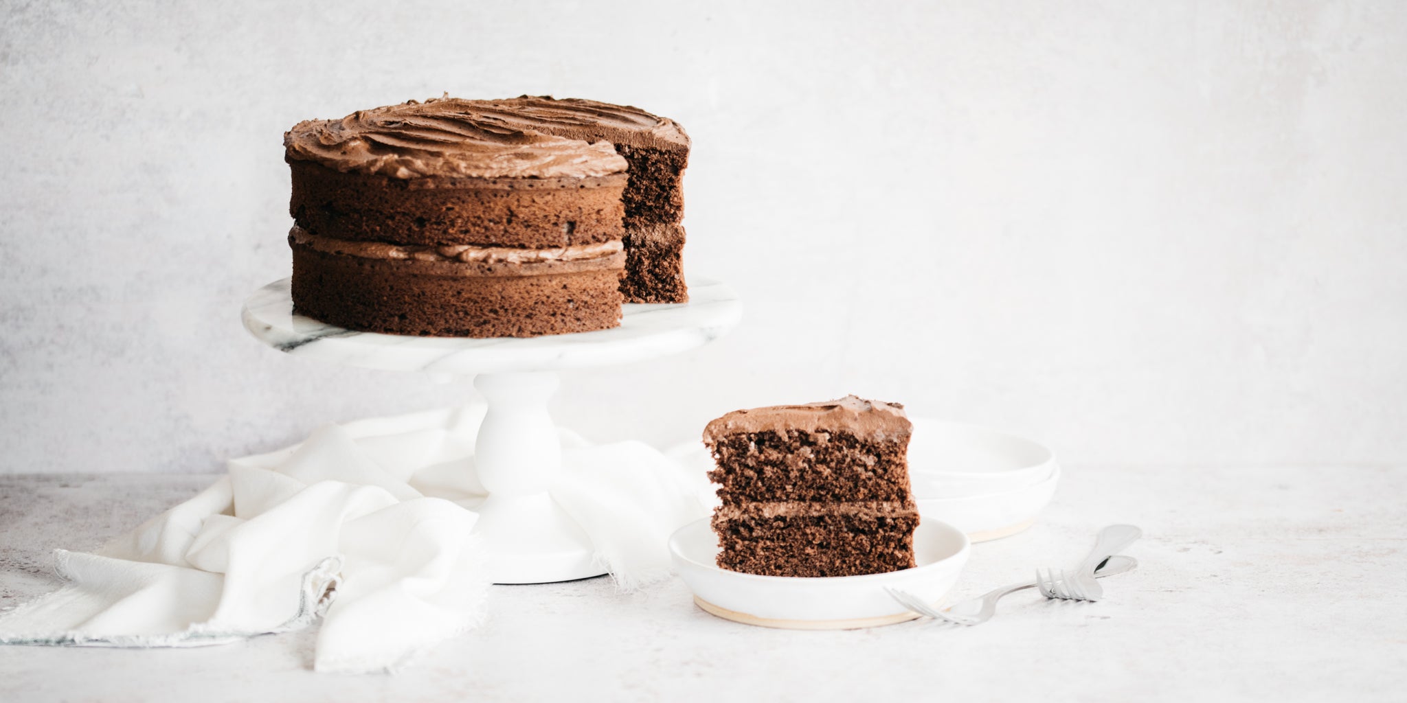 Chocolate cake topped with buttercream on a white cake stand with a slice removed on a white plate infront