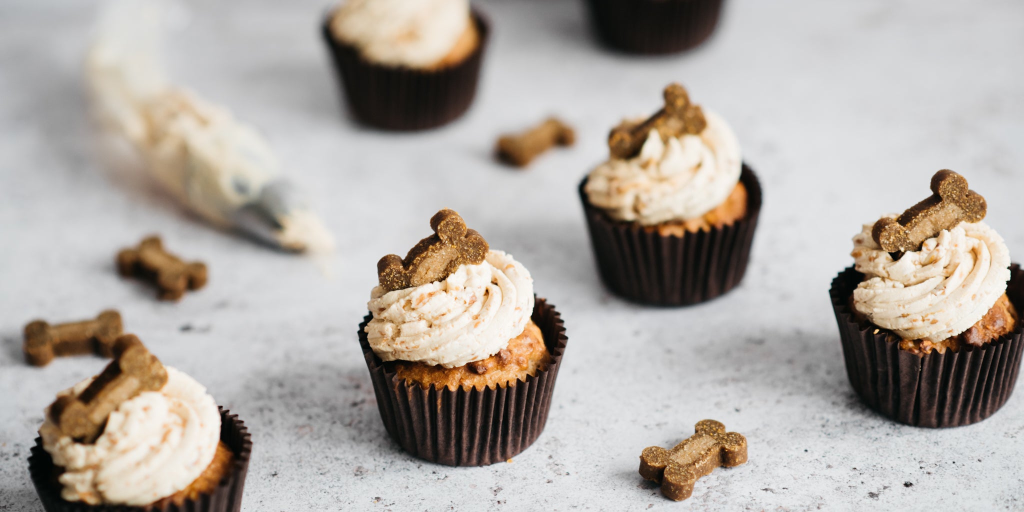 4 cupcakes in brown cases with frosting and dog treat toppings. Dog treats in the shape of bone scattered around base and a piping bag of frosting in the background