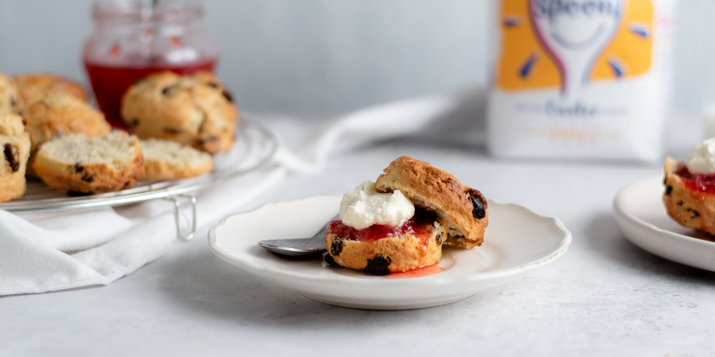 Close up of a Fruit Scone sliced in half, with a dollop of cream and jam. Bag of Silver Spoon Caster Sugar in the background