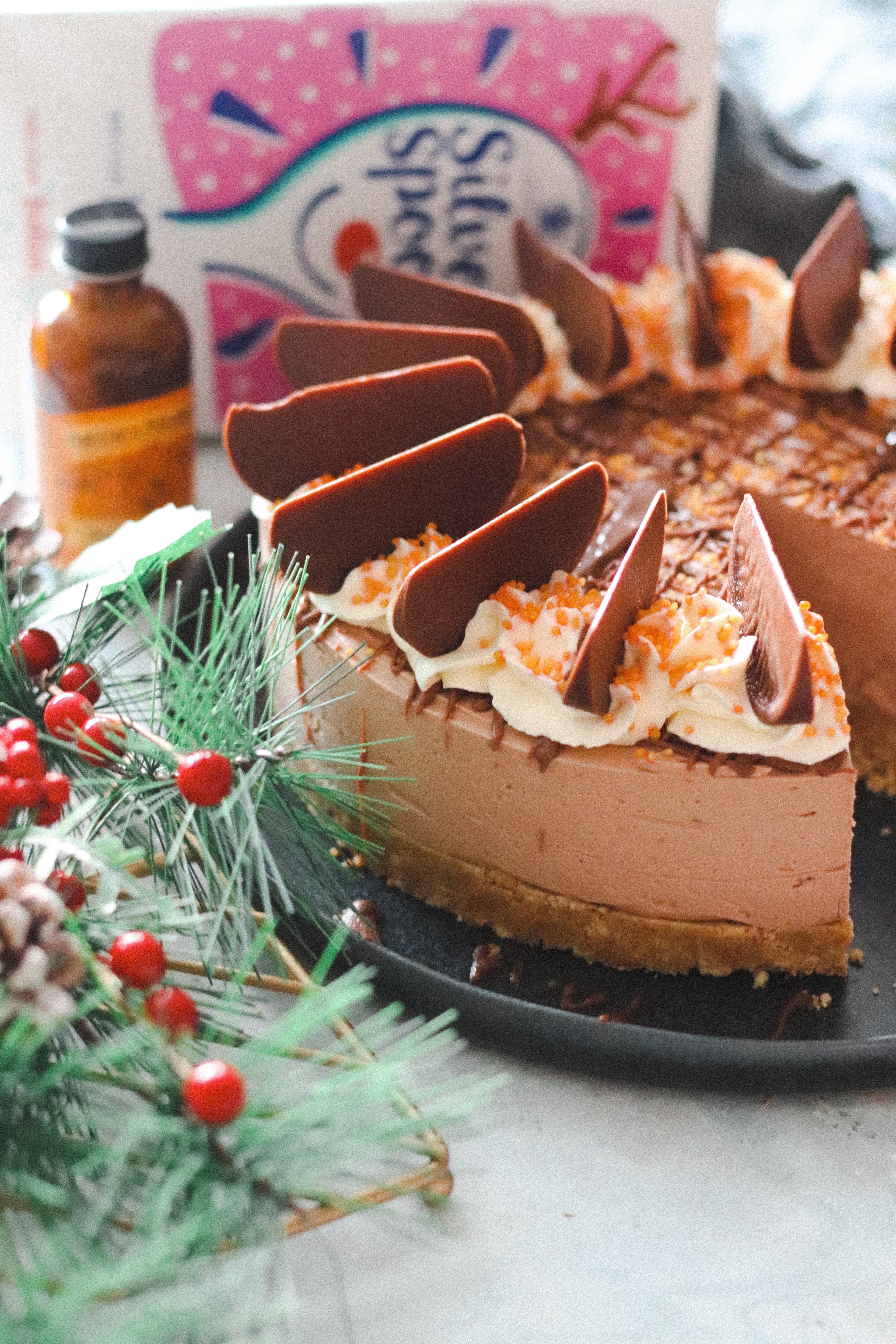 Close up of Chocolate Orange Cheesecake decorated with chocolate orange segments next to pine leaves, a box of Silver Spoon Icing Sugar and Nielsen-Massey Orange Extract