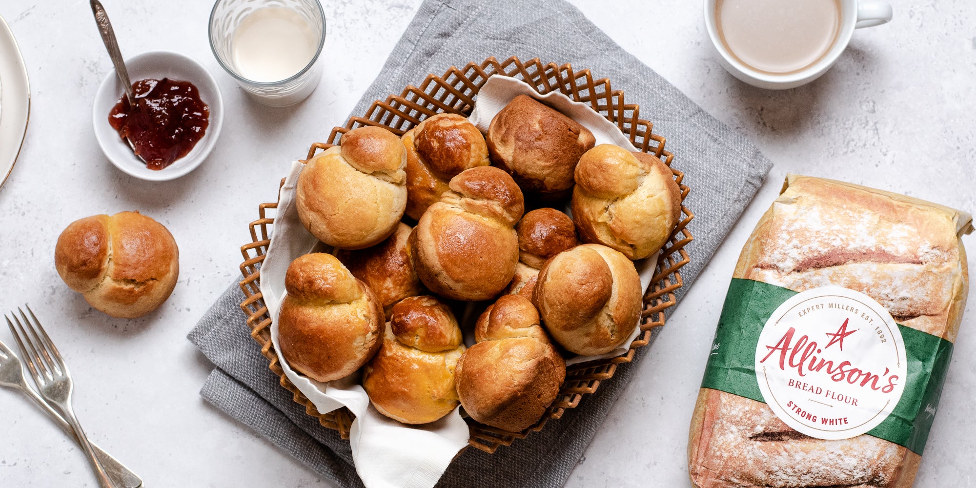 Top view of a batch of golden Brioche in a wicker basket. Next to a bag of Allinson's strong white flour, a small bowl of jam and a glass of milk