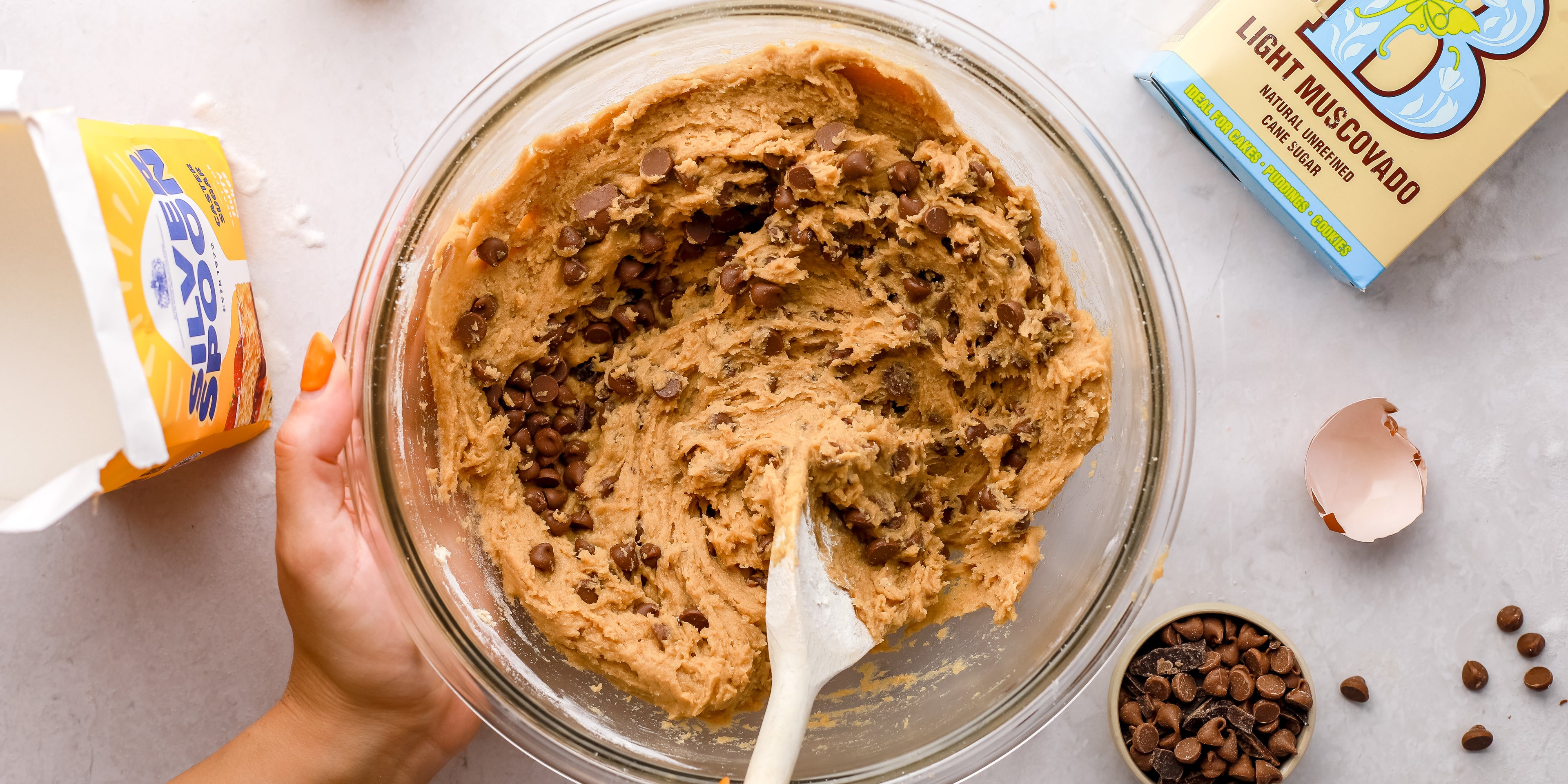 Cookie dough in a glass bowl with a wooden spoon and ingredients around the outside