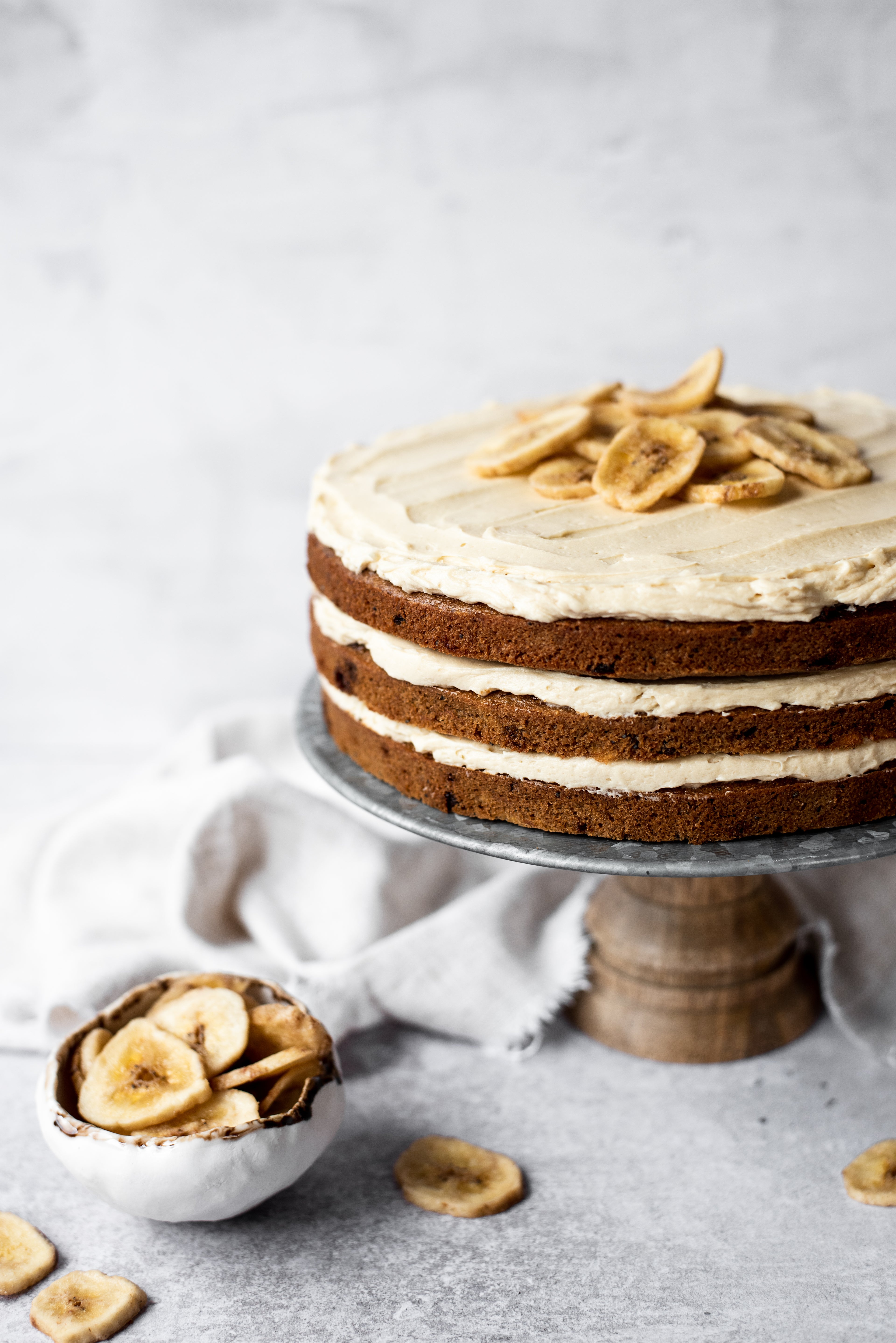 Banana Layer Cake with three layers on a cake stand with a bowl of banana chips 