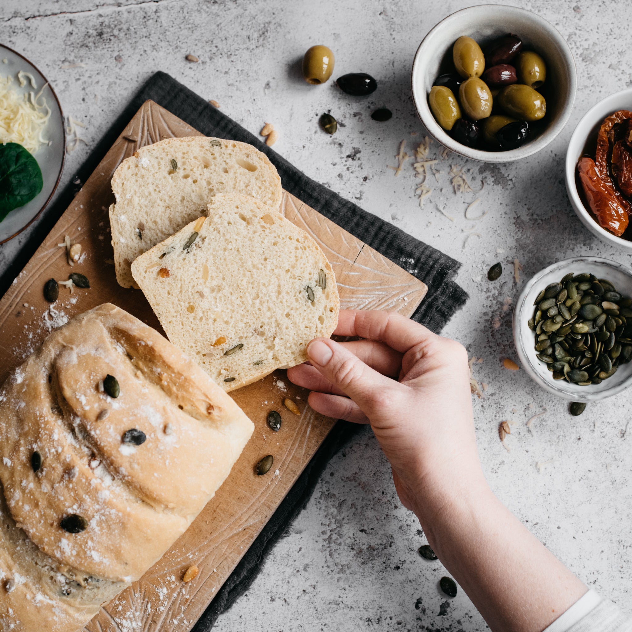 Spelt loaf with two slices a hand reaching in, seeds, olives and tomatoes in a bowl