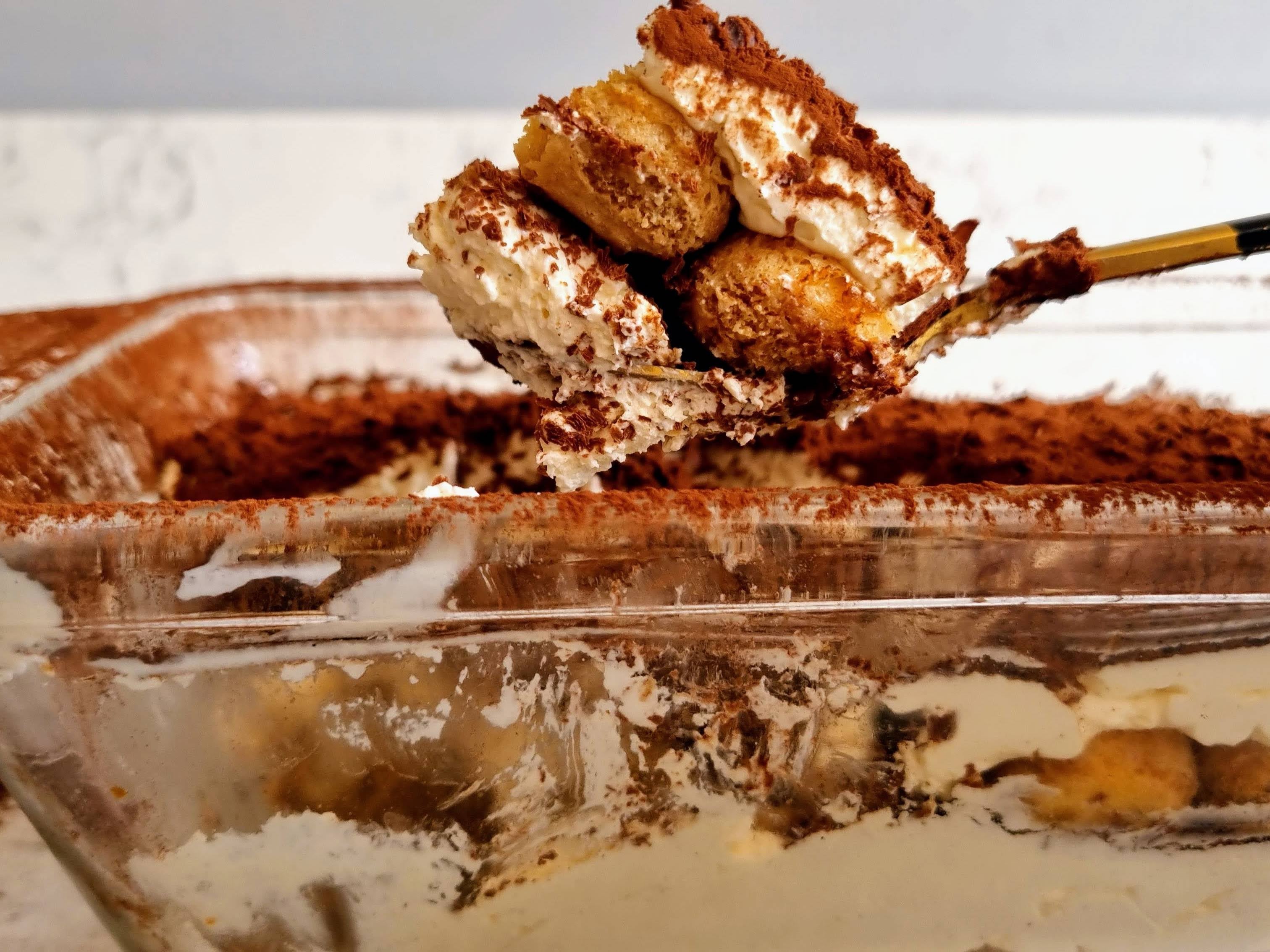 Homemade tiramisu with sponge fingers, cream and cocoa powder being taken out of baking dish with a spatula