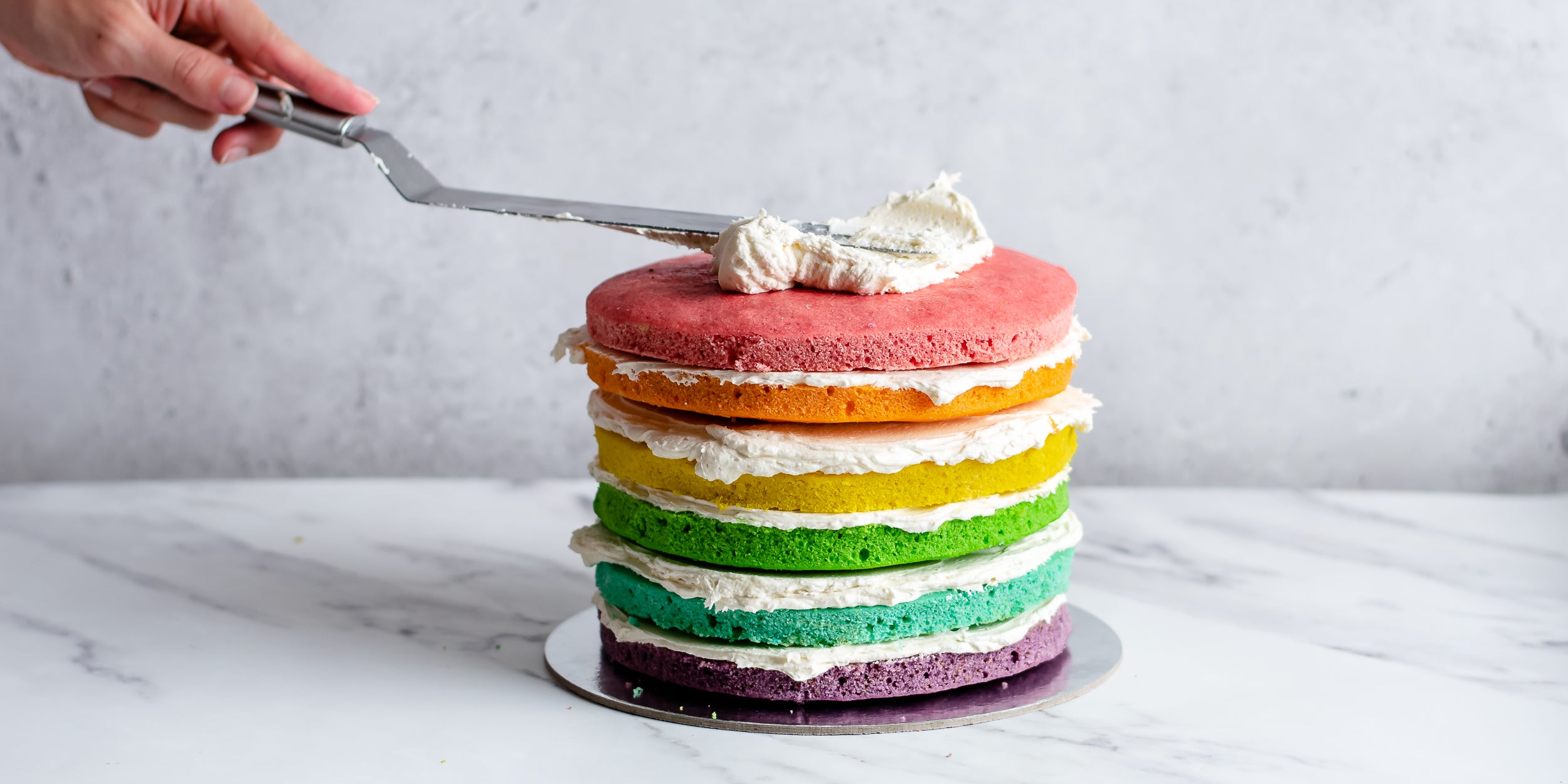 Hand reaching in with palette knife spreading buttercream on top of a pile of 6 rainbow colours sponge cakes