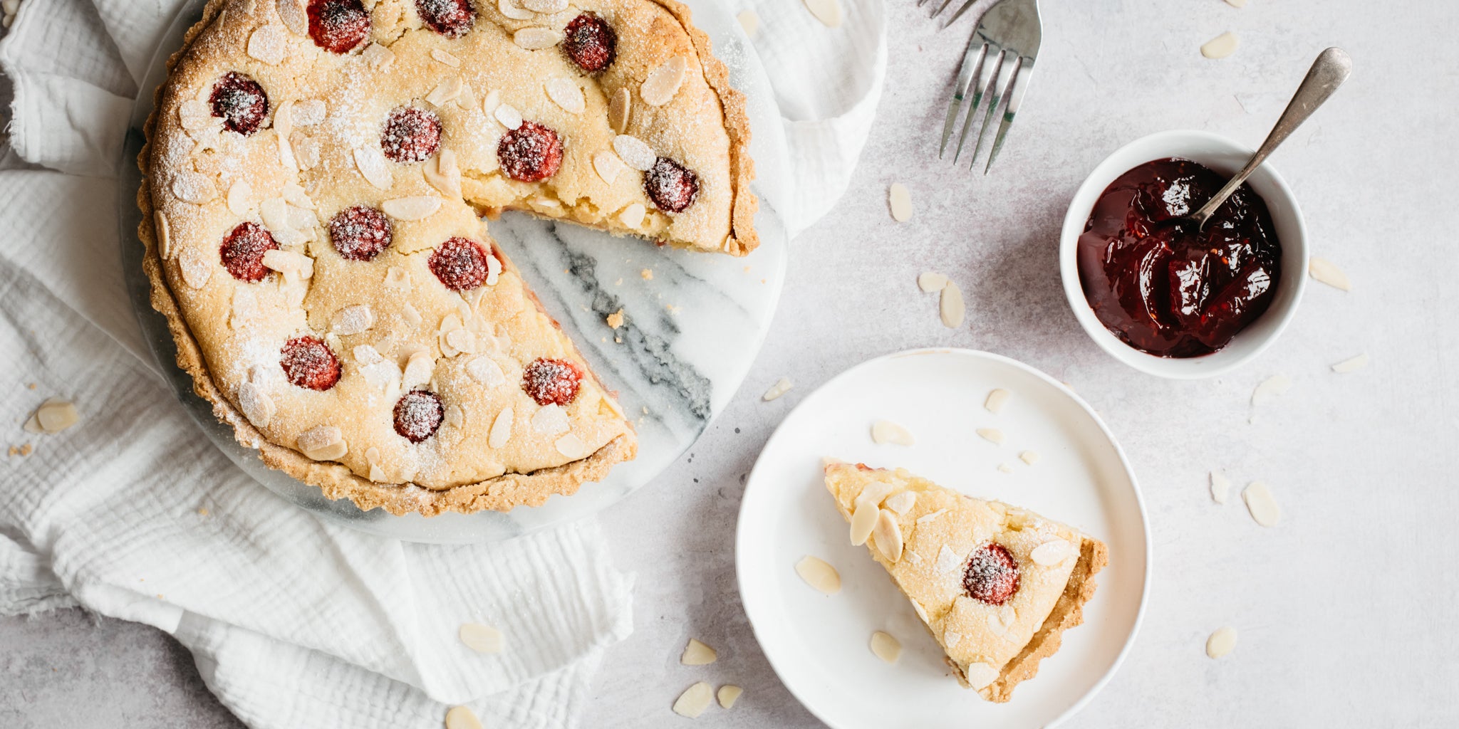 Shot of bakewell tart with slice removed. Slice on a white plate in front beside a pot of jam with a spoon