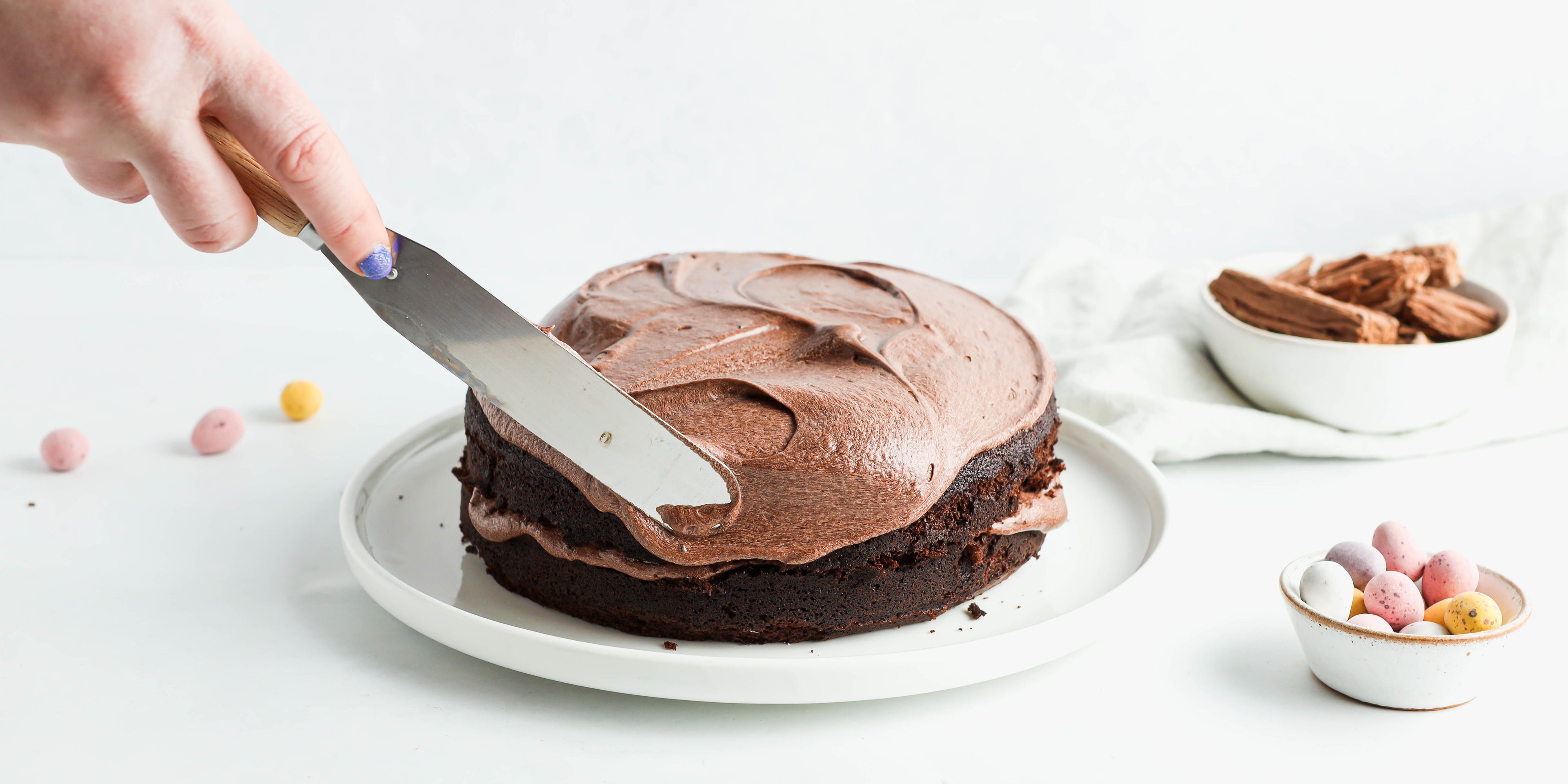 Close up of a Chocolate Nest Cake being hand decorated with a palette knife ready to top with mini eggs and flake