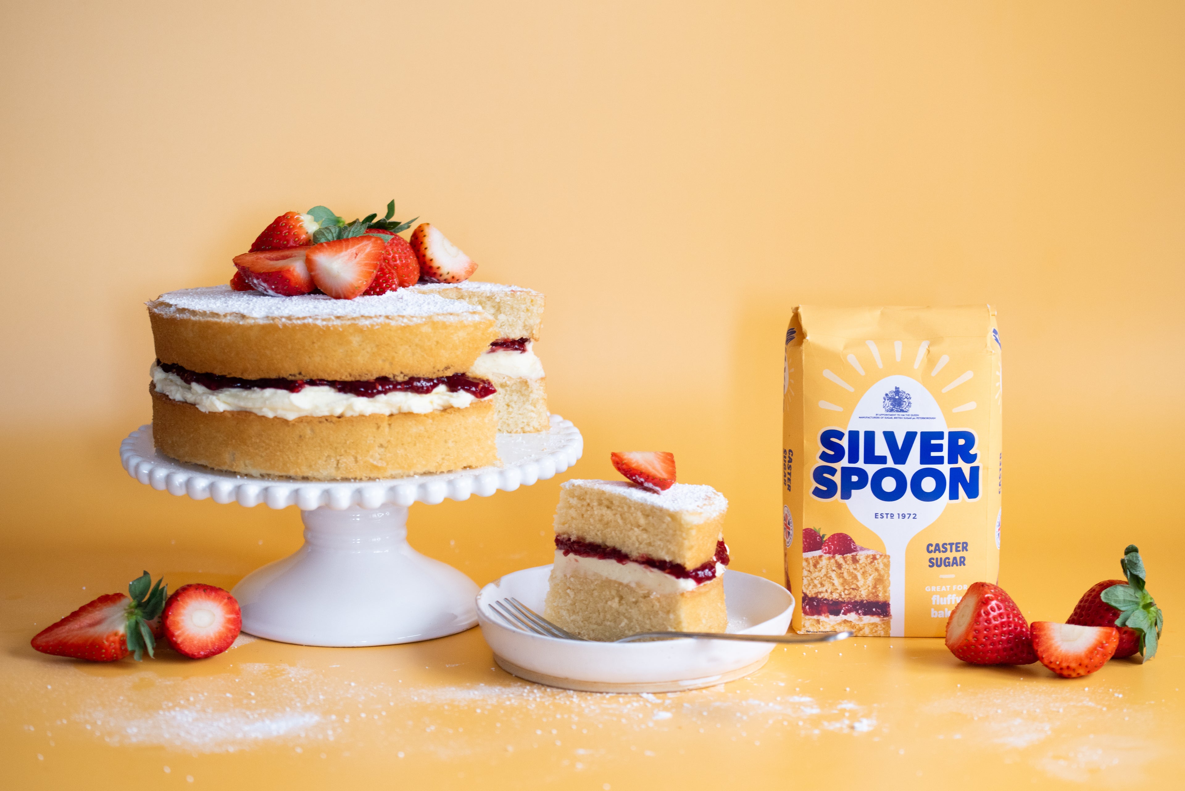 Victoria sponge on a white cake stand with a slice on a plate beside it. There is a pack of Silver Spoon sugar next to it. 