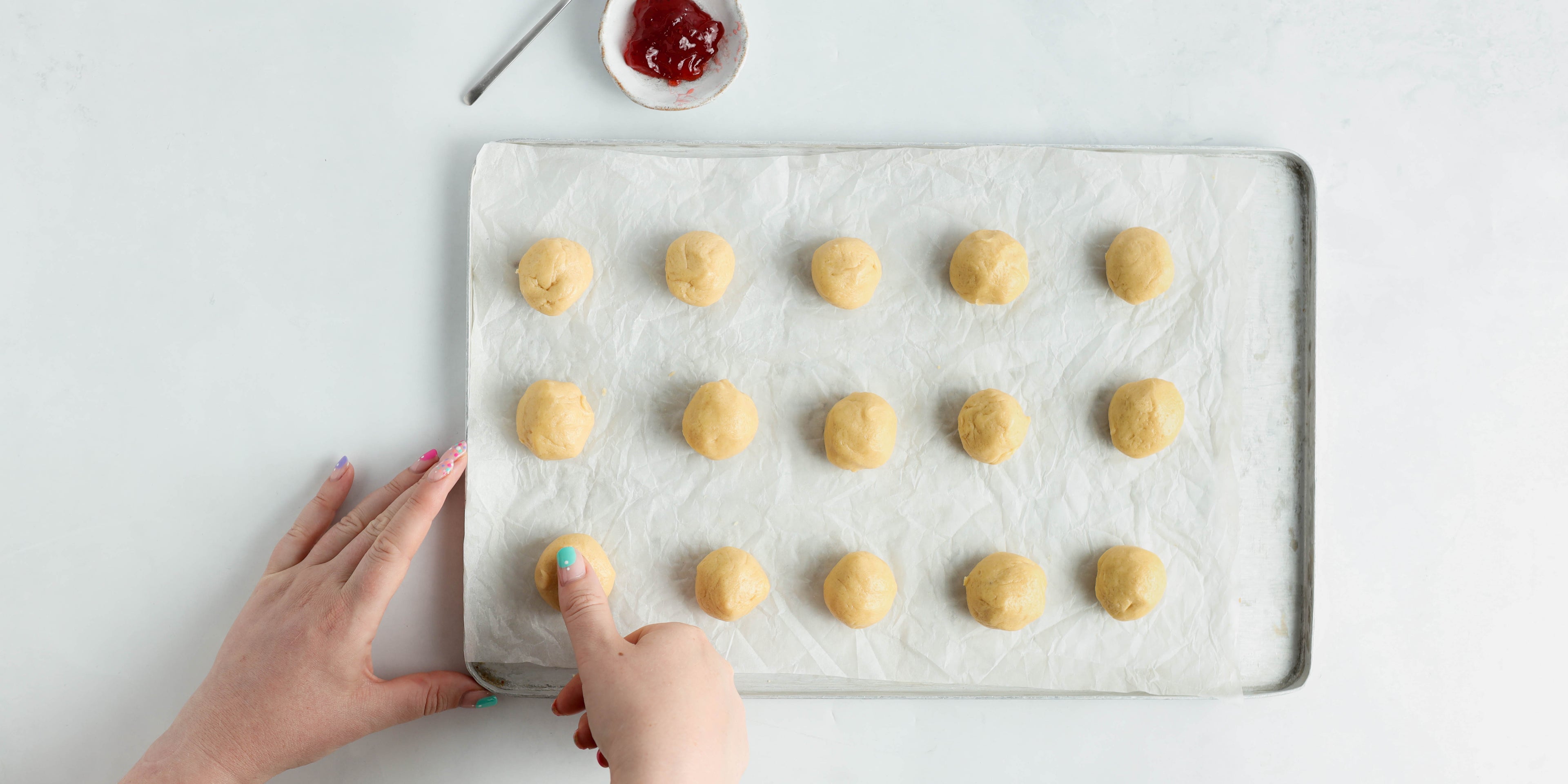 15 balls of cookie dough on a tray with a thumb pushing into one. A pot of jam at the top