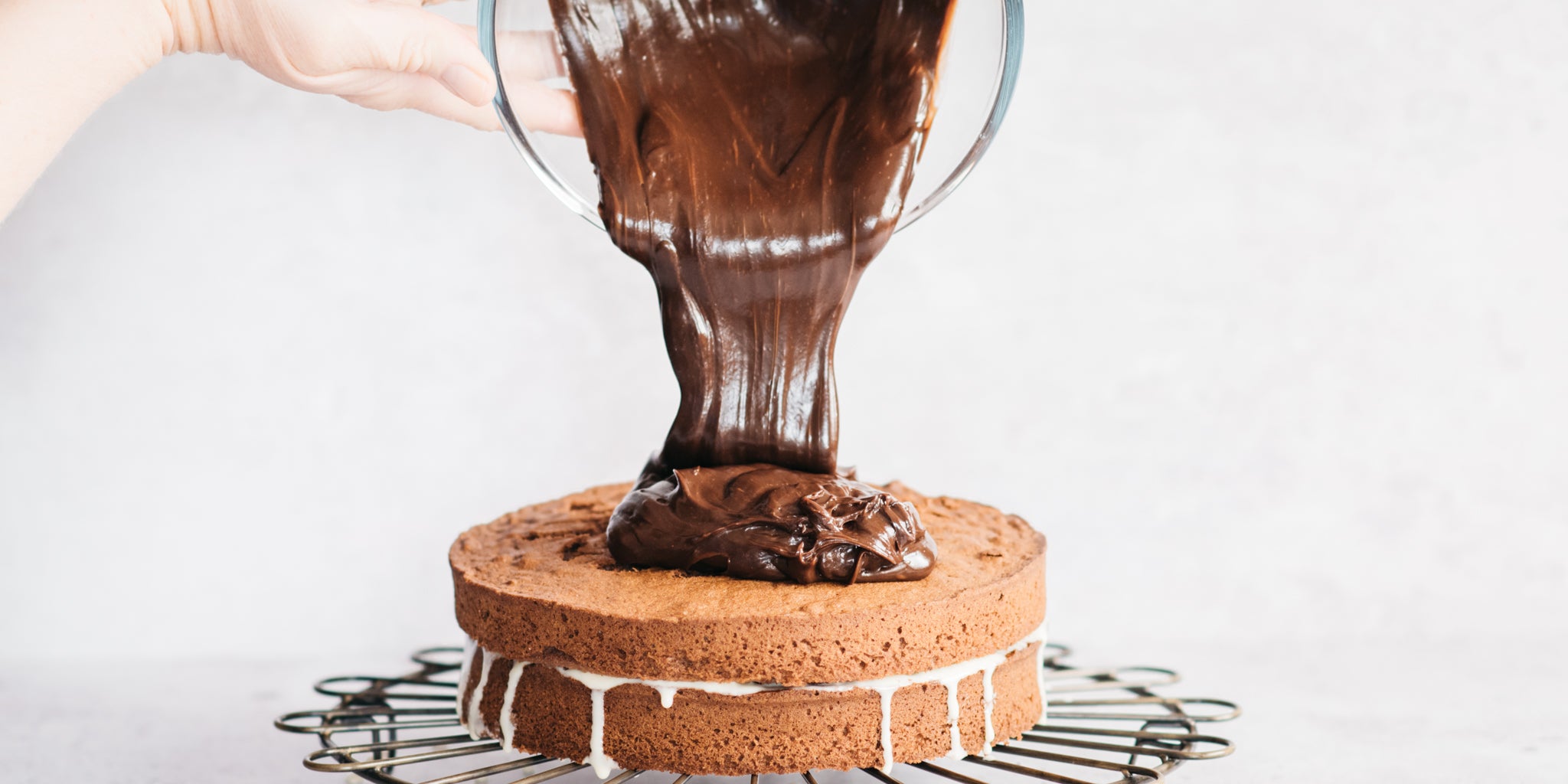 Bowl of melted chocolate being poured on top of sponge cake on cooling rack
