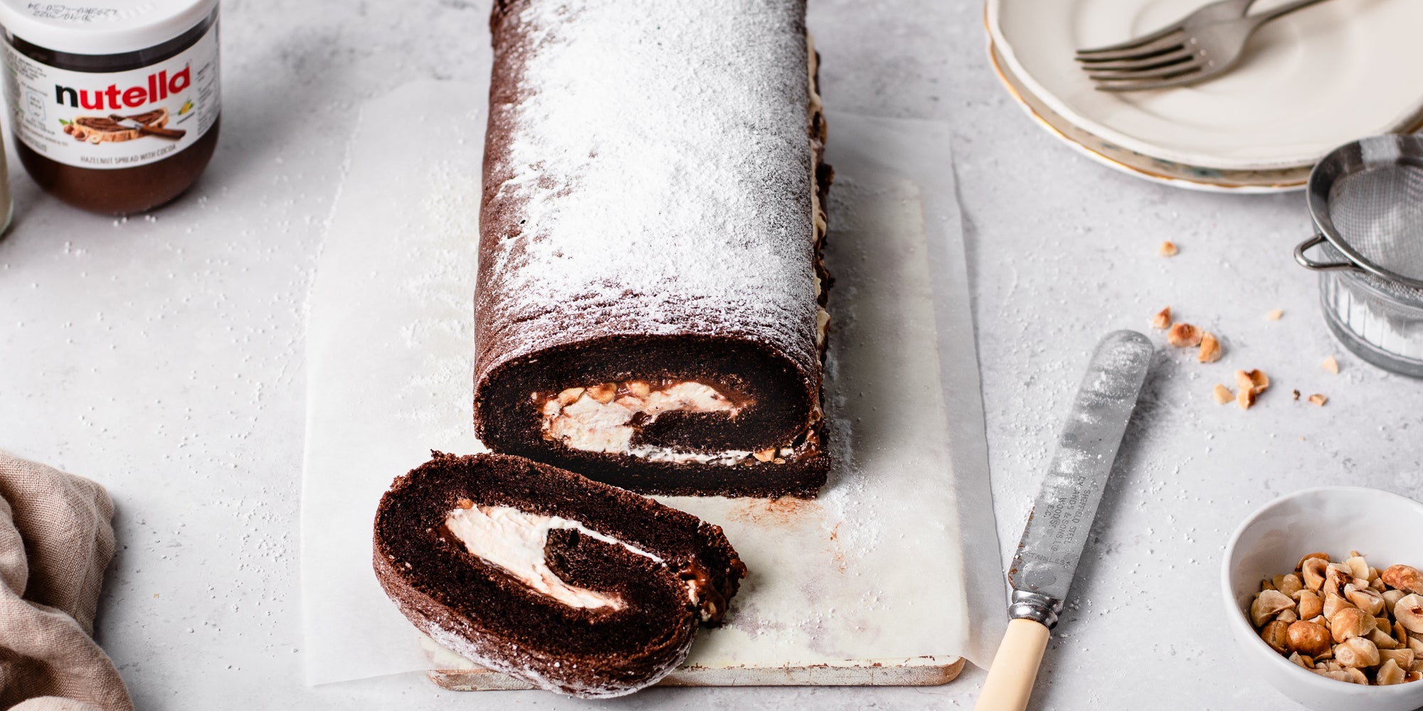 Top view of Chocolate Roulade on a serving board, next to a knife, a bowl of chopped hazelnuts and plates with forks ready to serve