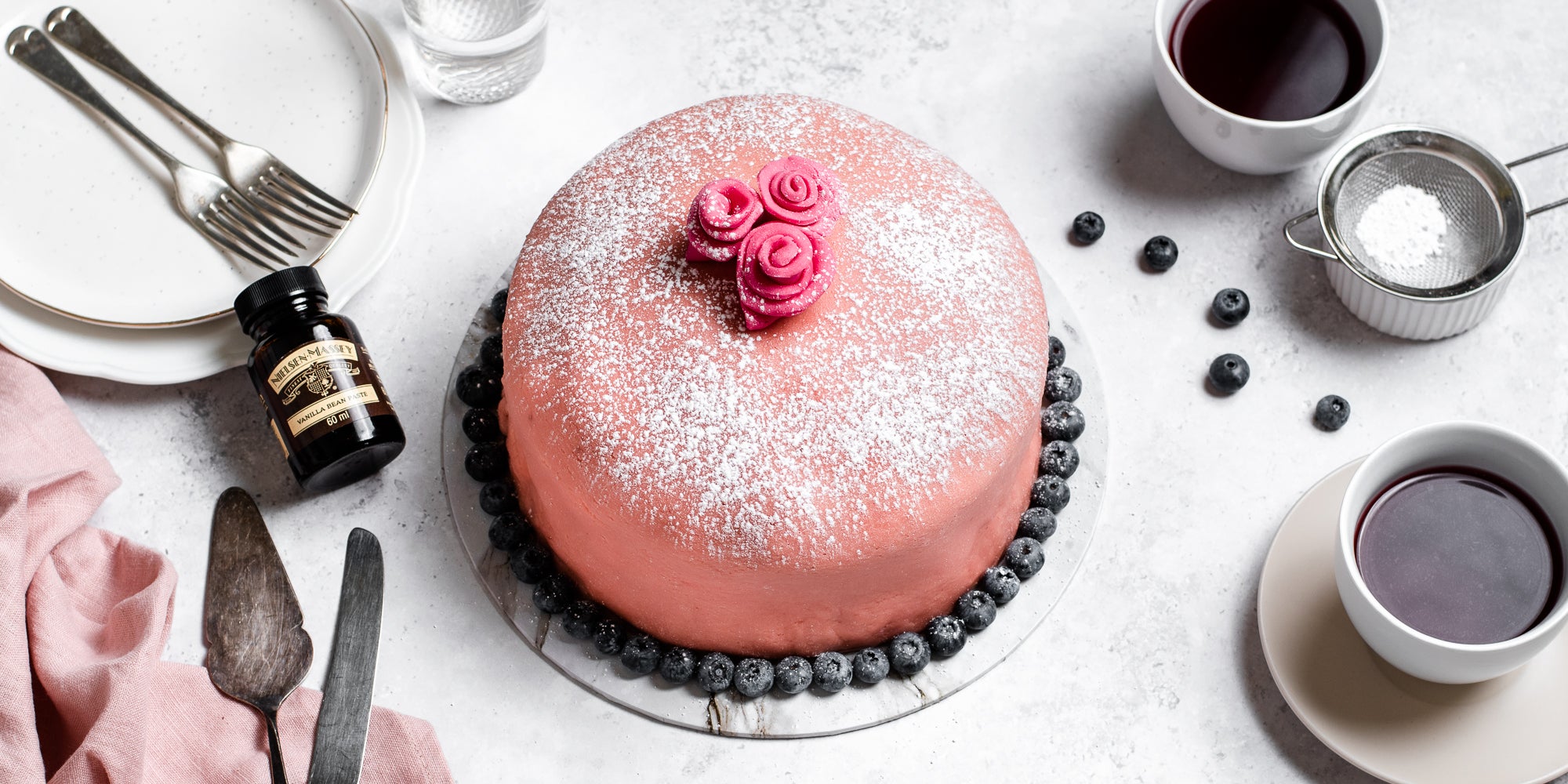 Top view of Prinsesstarta dusted with icing sugar and decorated with blueberries. Bottle of Nielsen-Massey Vanilla Extract next to the Prinsesstarta