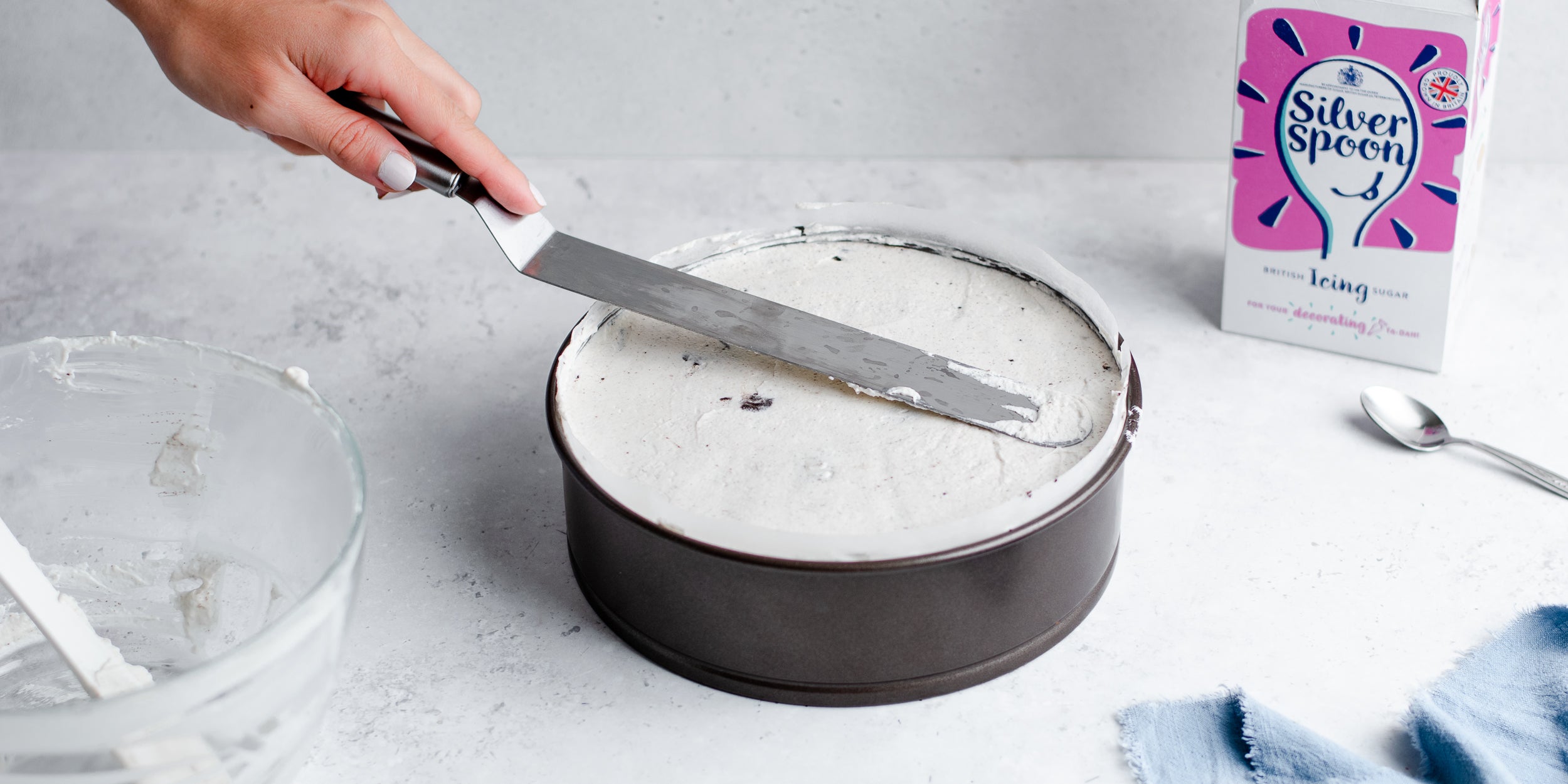 Circular baking tin with hand smoothing down mixture using a pallet knife. Sugar pack beside it