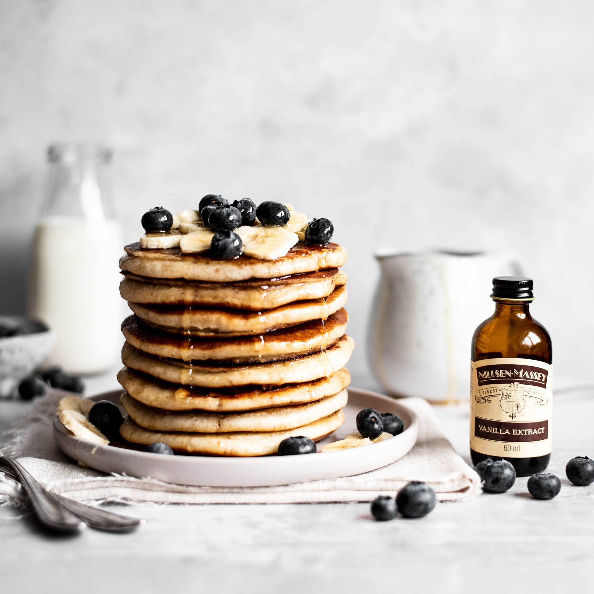 A stack of vegan pancakes with blueberries and sliced banana on top next to a bottle of Nielsen Massey vanilla extract