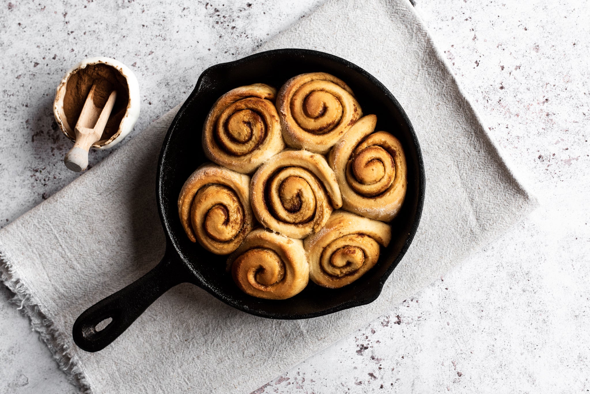 Uniced cinnamon rolls in a skiller with a pot of cinnamon beside it