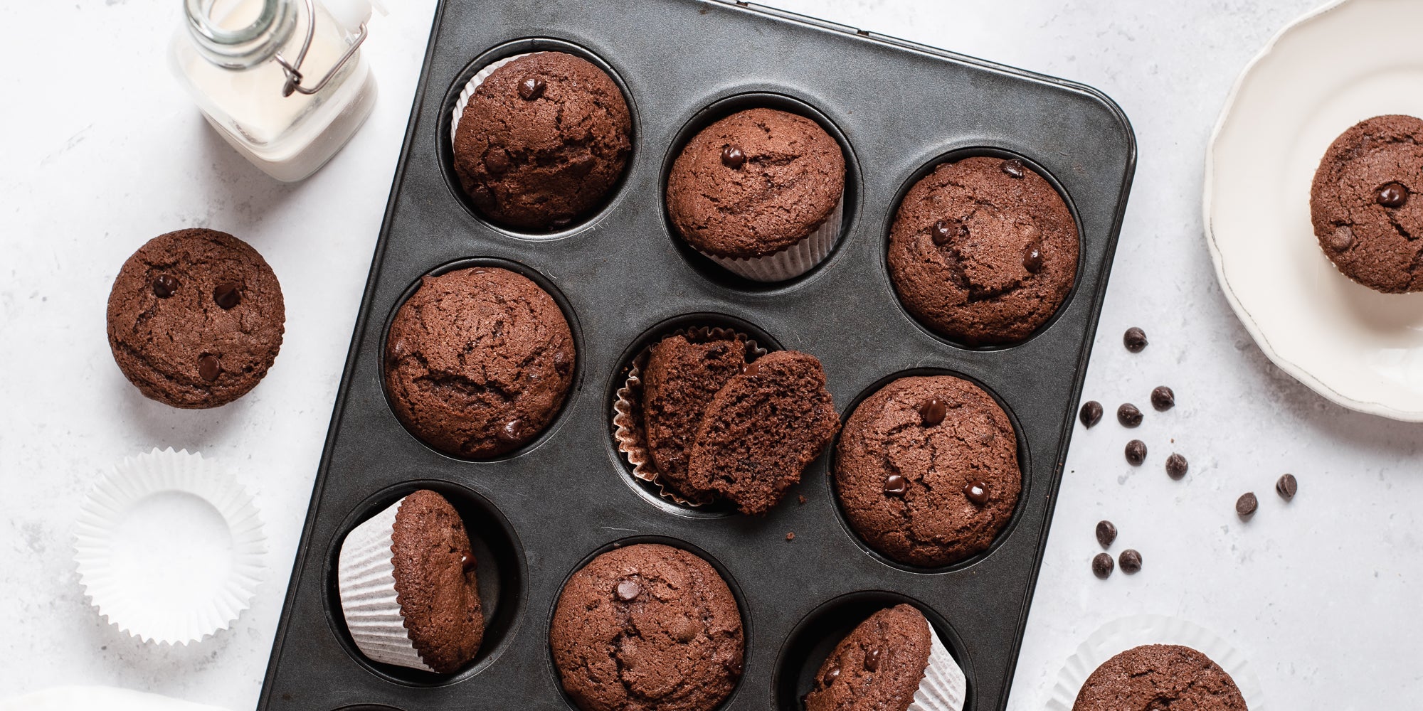Gluten Free Chocolate Muffins in a muffin tray with chocolate chips and a glass bottle of milk