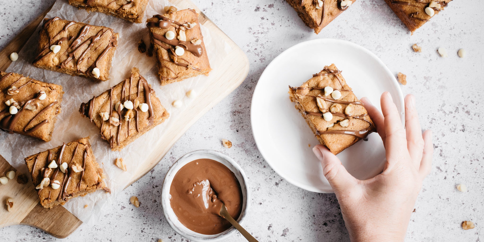 Overhead shot of peanut butter blondies cut into squares. One on a white plate with a hand reaching in. Pot of melted chocolate and a spoon