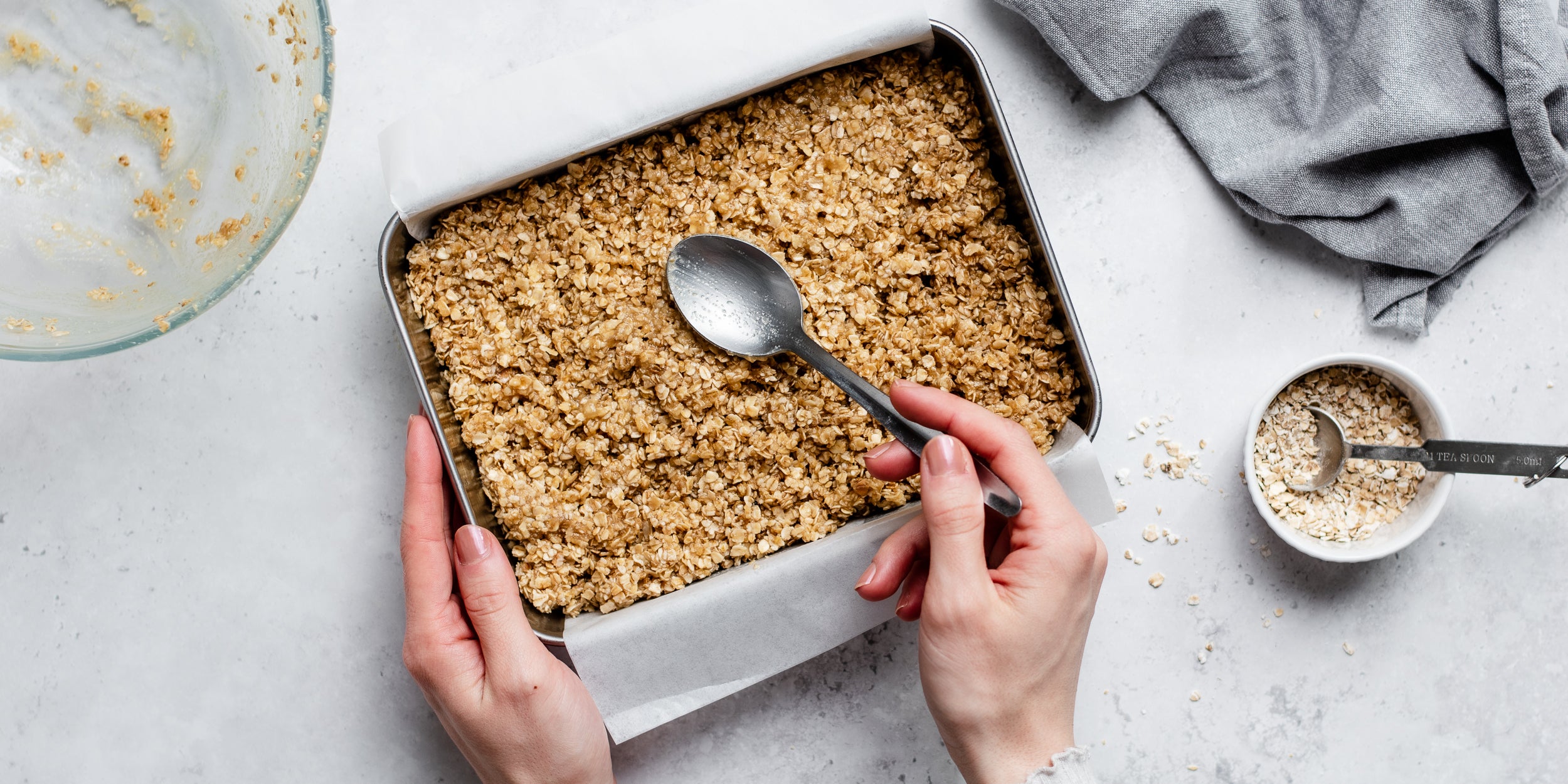 Easy Flapjack mix being pressed into a lined baking tray, with a hand holding a spoon, next to a ramekin of oats 