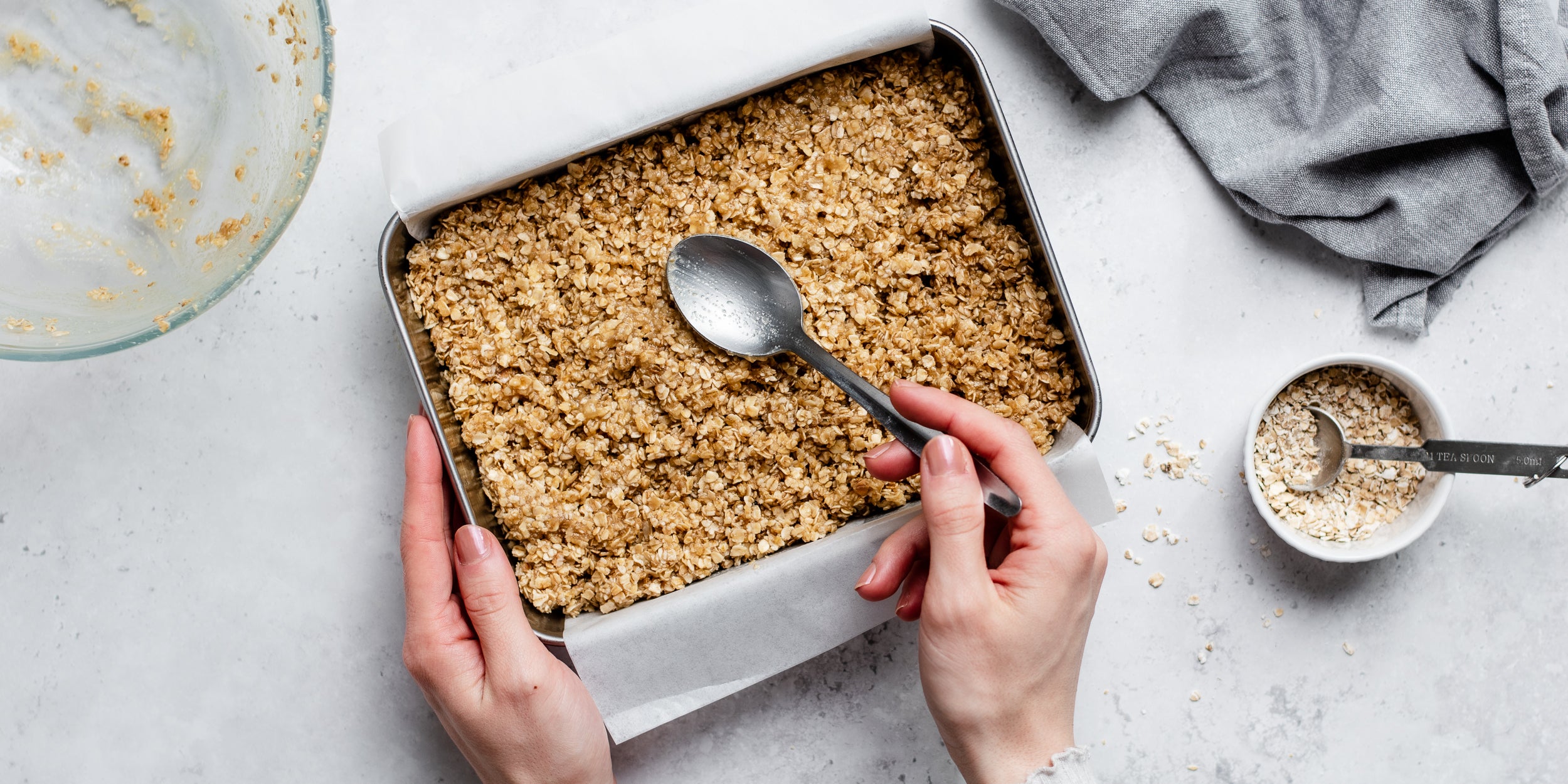 Easy Flapjack mix being pressed into a lined baking tray, with a hand holding a spoon, next to a ramekin of oats