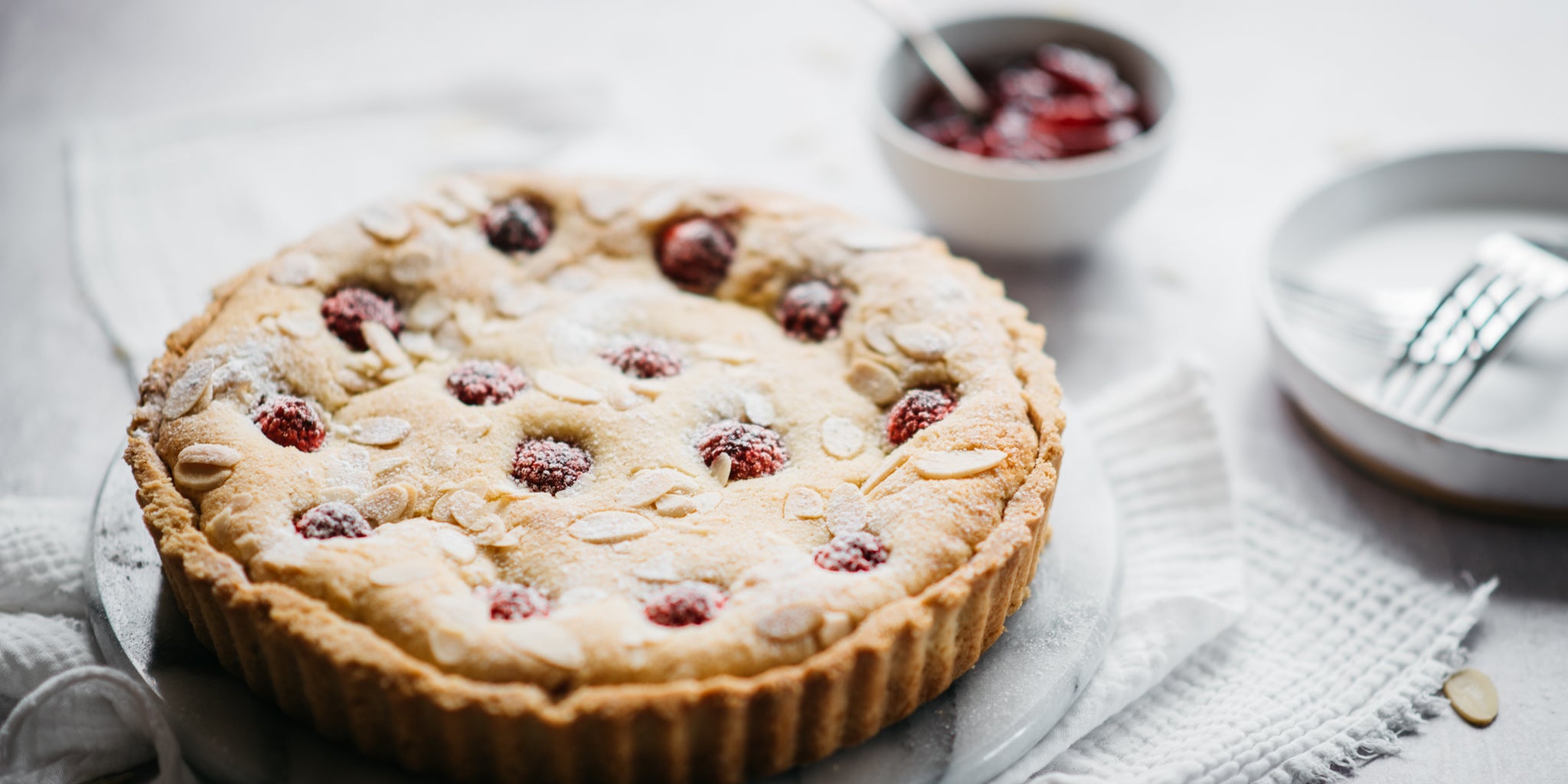 Close up of a bakewell tart on a white plate with a pot of jam and empty white plate in background