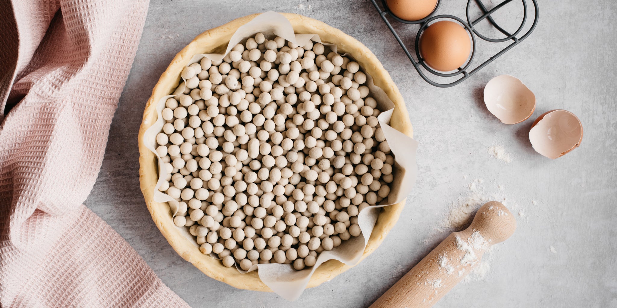 Simple Bakewell Tart pastry being blind baked with pastry beans, next to a rolling pin and egg shells