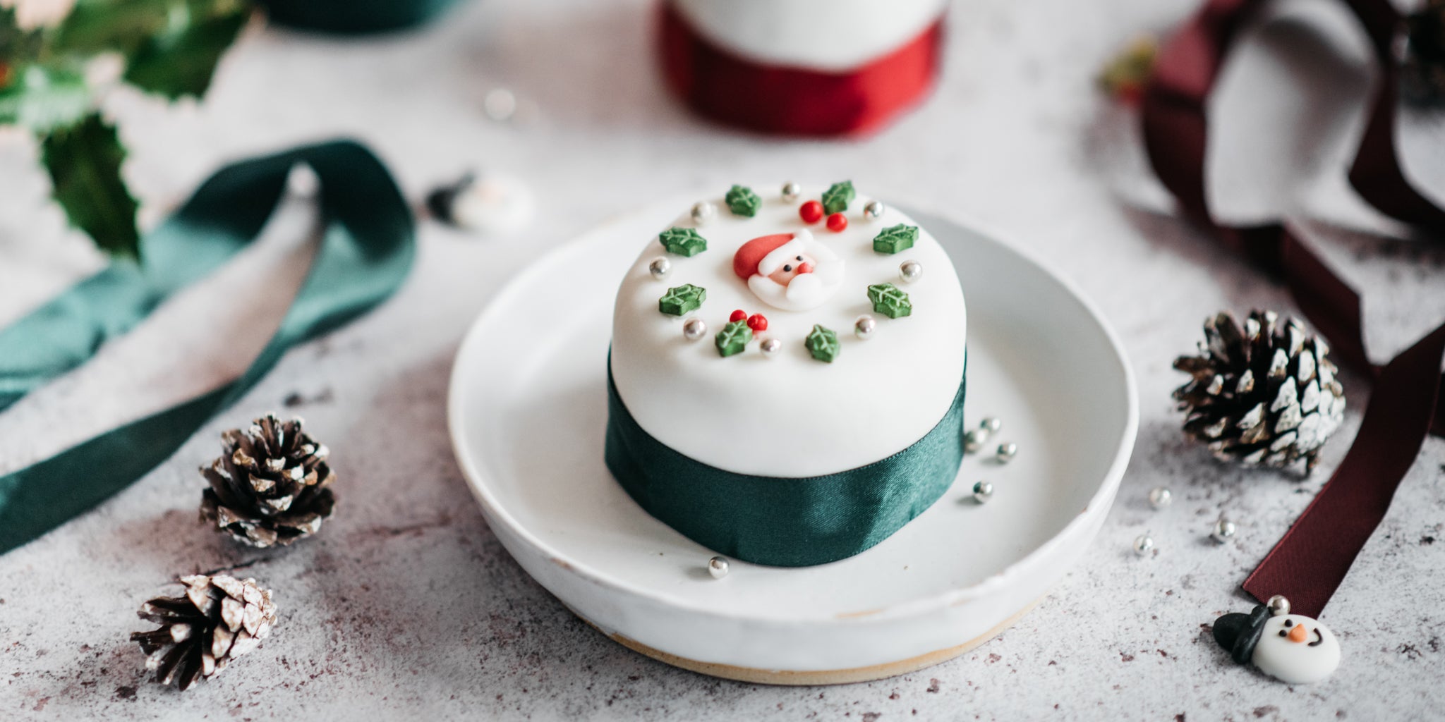 Close up of a mini christmas cake decorated with green ribbon and a santa decoration. Surrounded by ribbon, holly and pinecones