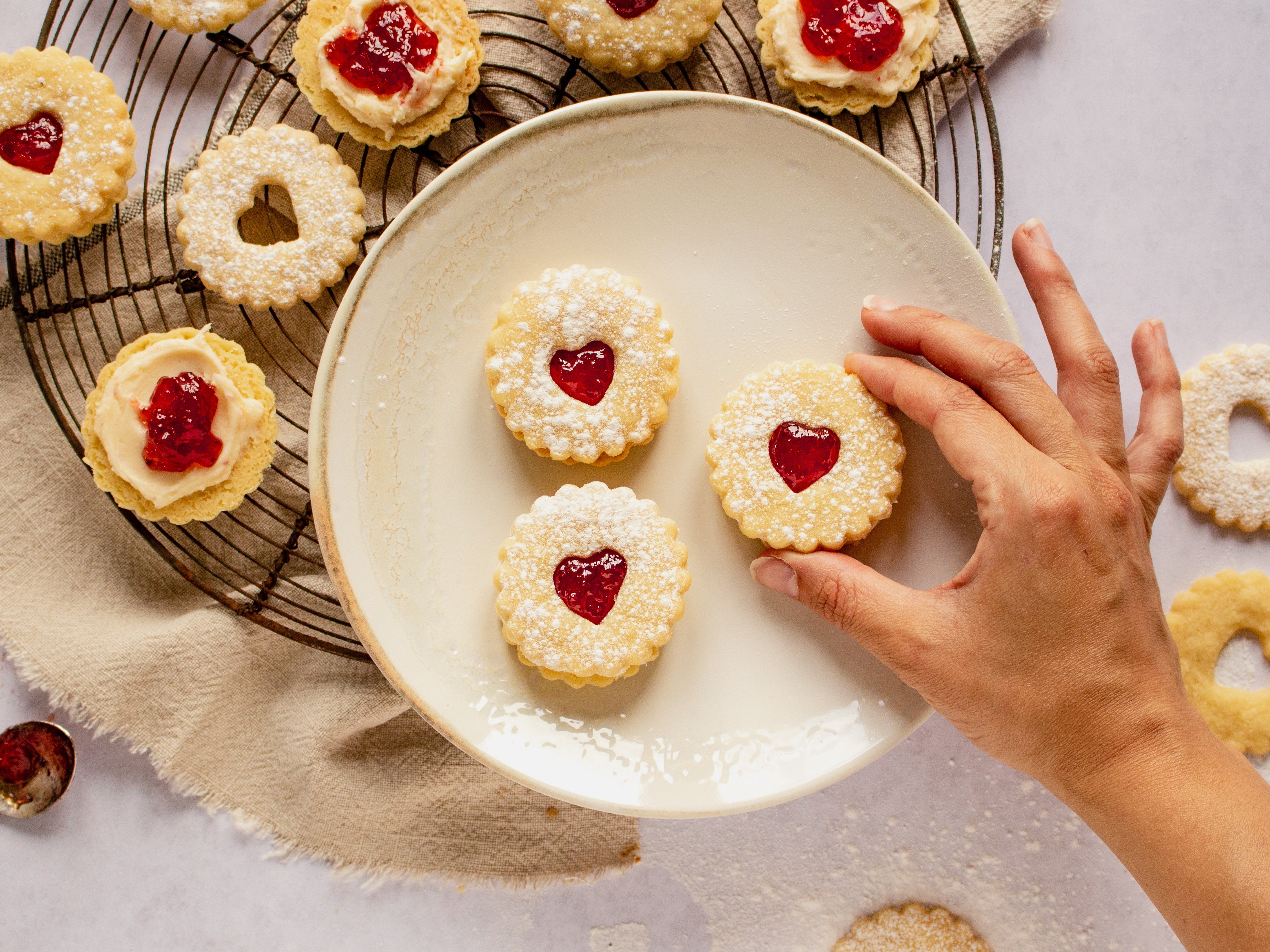 3 Jammy Dodgers on white plate with hand reaching in. Pulled apart biscuits on cooling rack