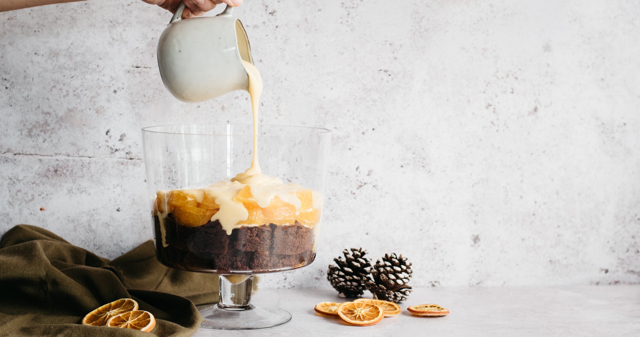 Chocolate Orange Trifle being layered with Baileys custard ontop of the base of chocolate sponge, hand holding jug drizzling the custard. 