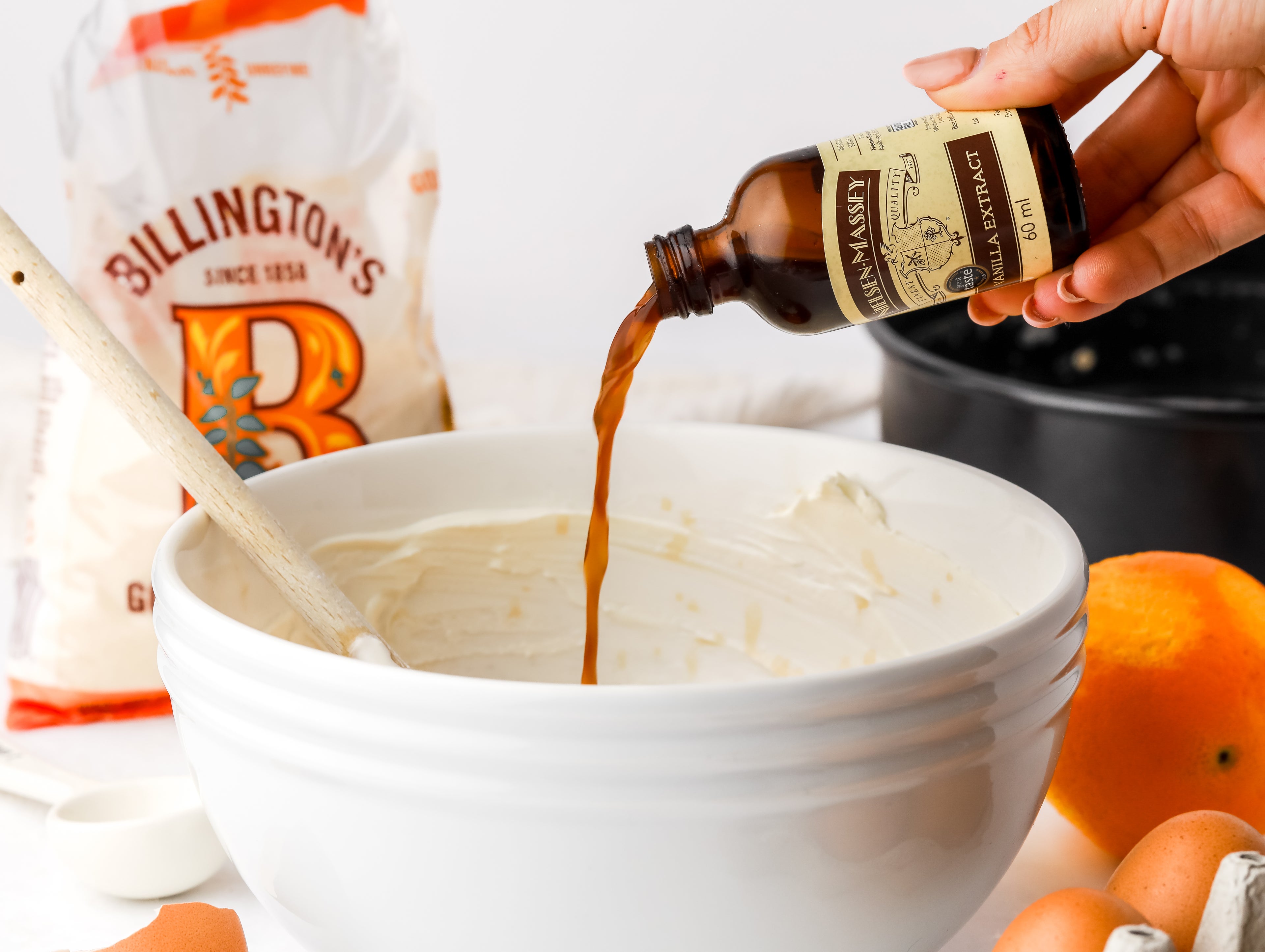 Vanilla extract being poured into a bowl of cheesecake mix with wooden spoon in. Baking tin, orange and pack of sugar in background