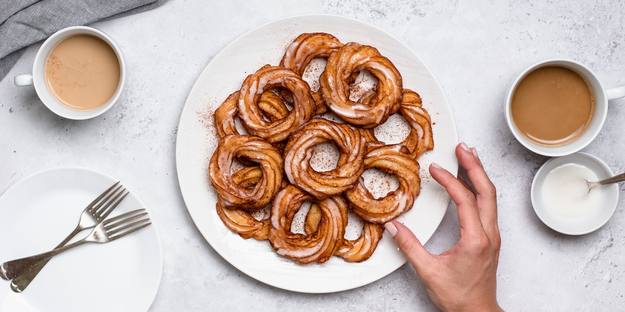 Top view of a plate of Apple Cider Crullers, with a hand reaching to pick one. Next to cups of tea, and a plate with forks ready to serve