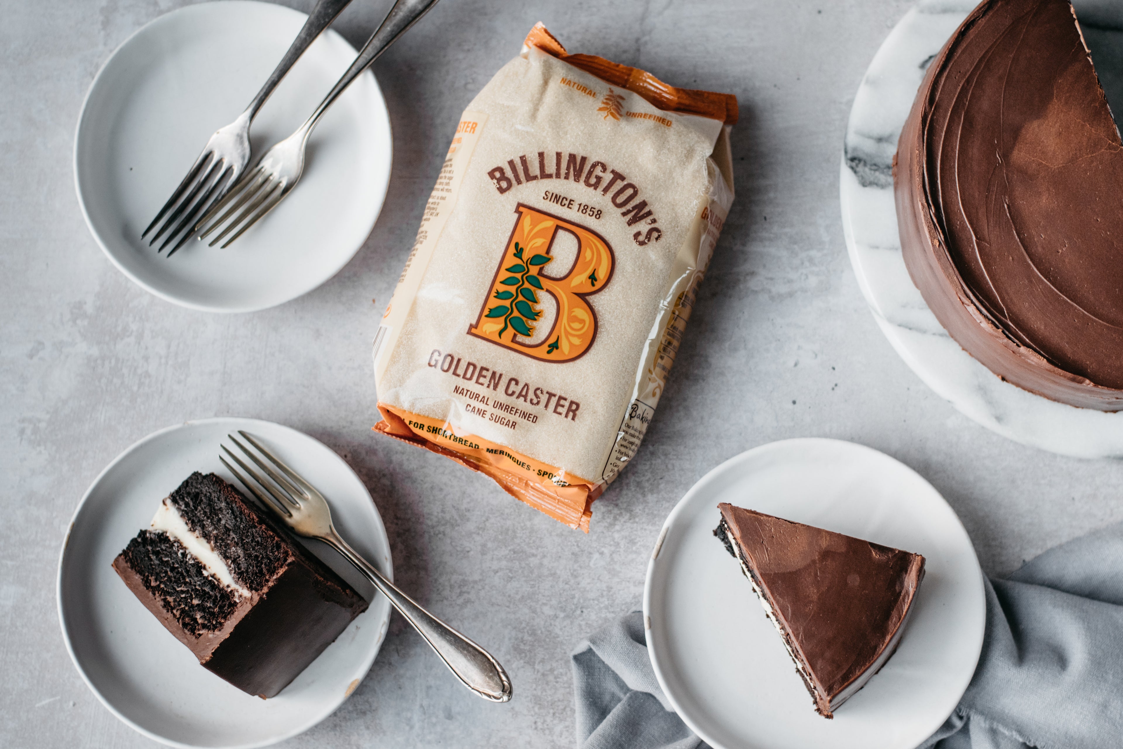 Top view of slices of Best Chocolate Cake served on plates, next to a flat lay bag of Billington's Golden Caster Sugar