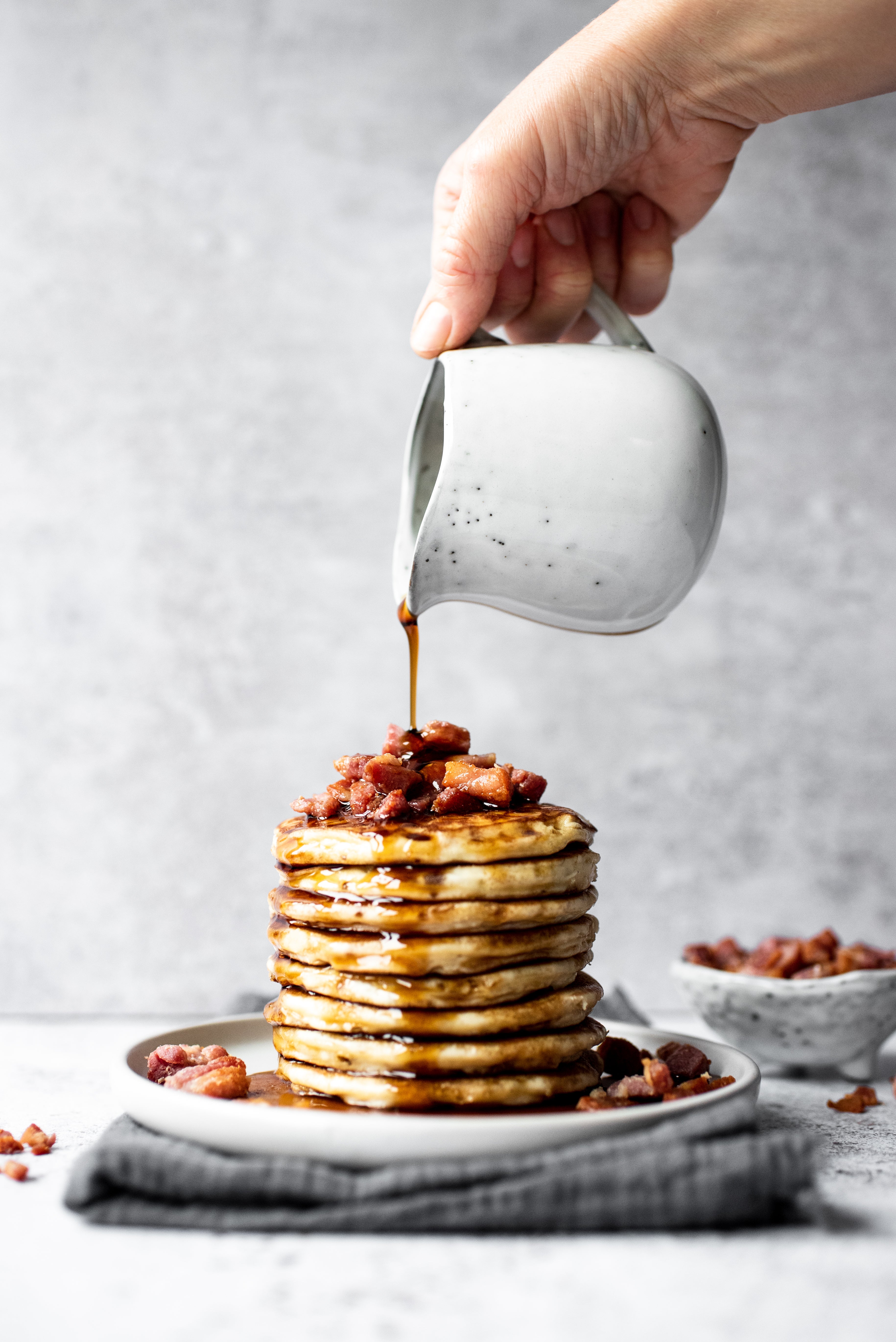 Stack of pancakes topped with bacon. Hand and jug pouring maple syrup on top