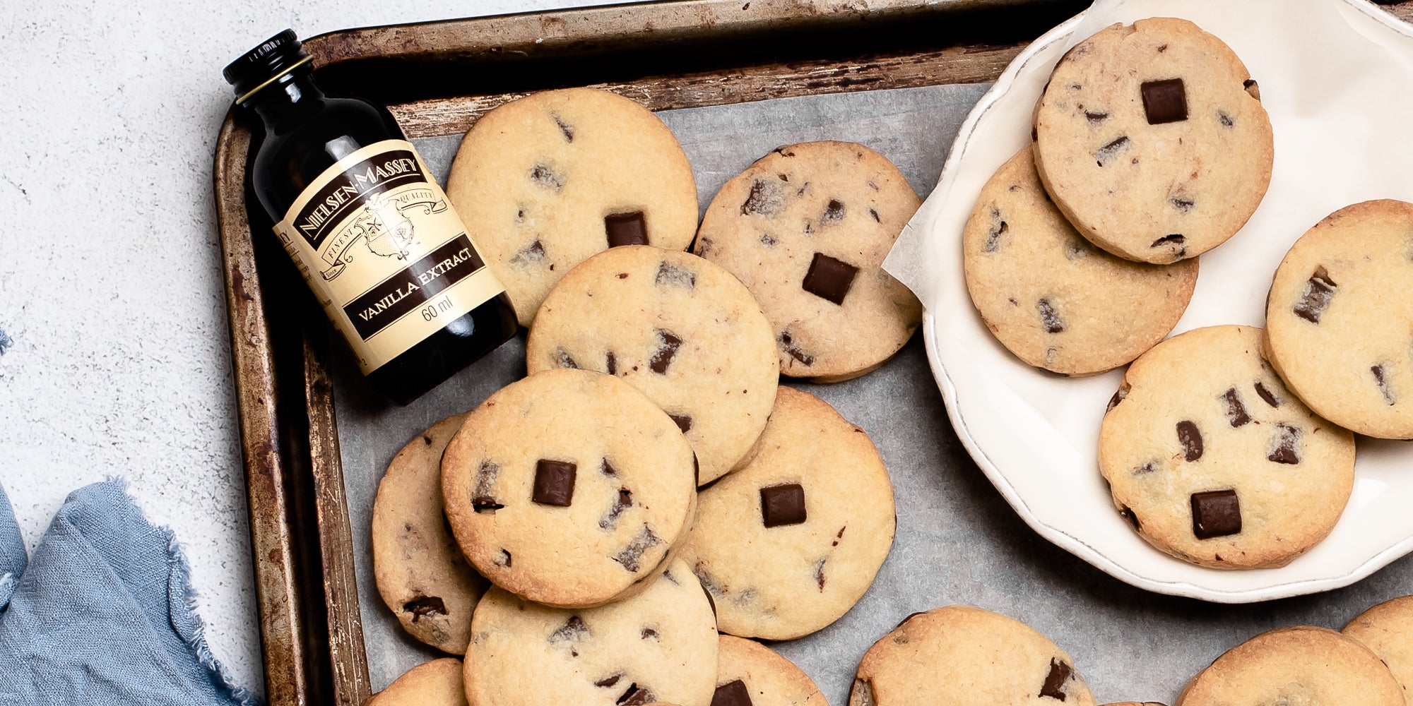 Batch of Chocolate Chunk Shortbread on baking paper on a baking tray, next to a bottle of Nielsen-Massey vanilla extract and a plate with 4 shortbread cookies on it