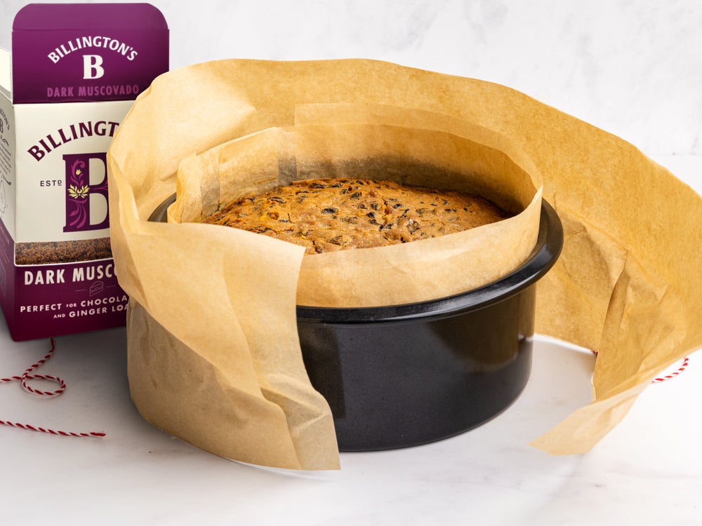 A pack of Billington's Dark Muscovado sugar next to a round freshly baked fruit cake thats wrapped in brown paper in the tin.