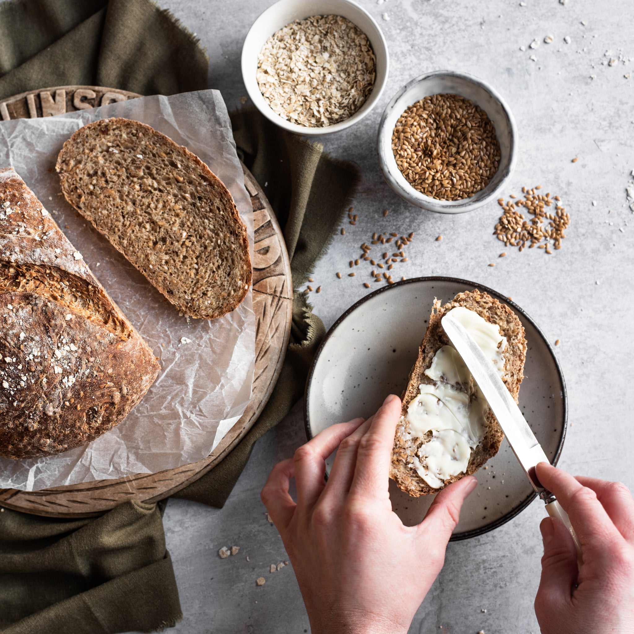 Overhead shot of round bread and one slice cut in front. Two bowls of seeds and hands buttering a slice of bread