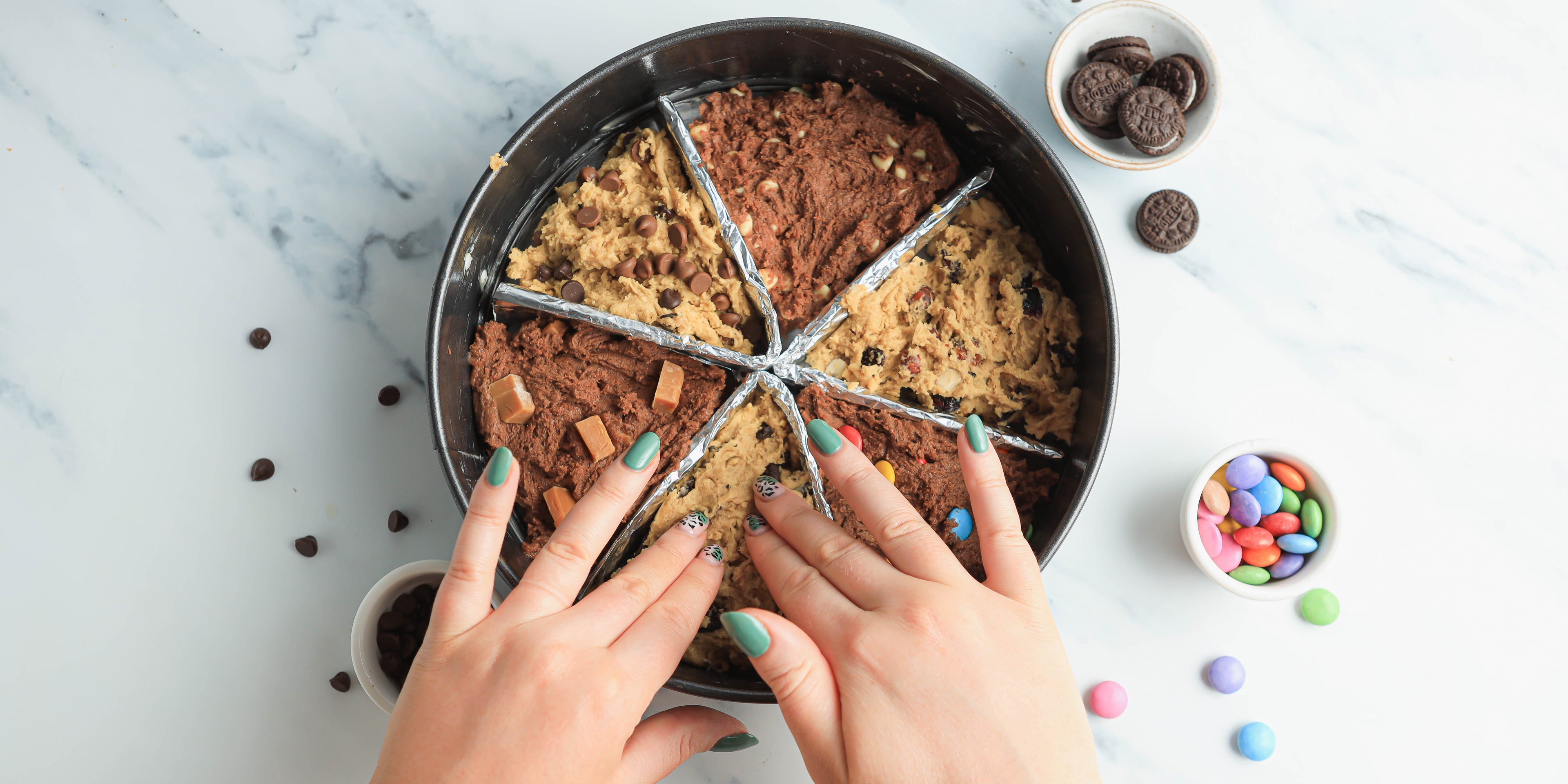 Two hands pushing down cookie dough mix into a circular cake tin with biscuits and sweets in background