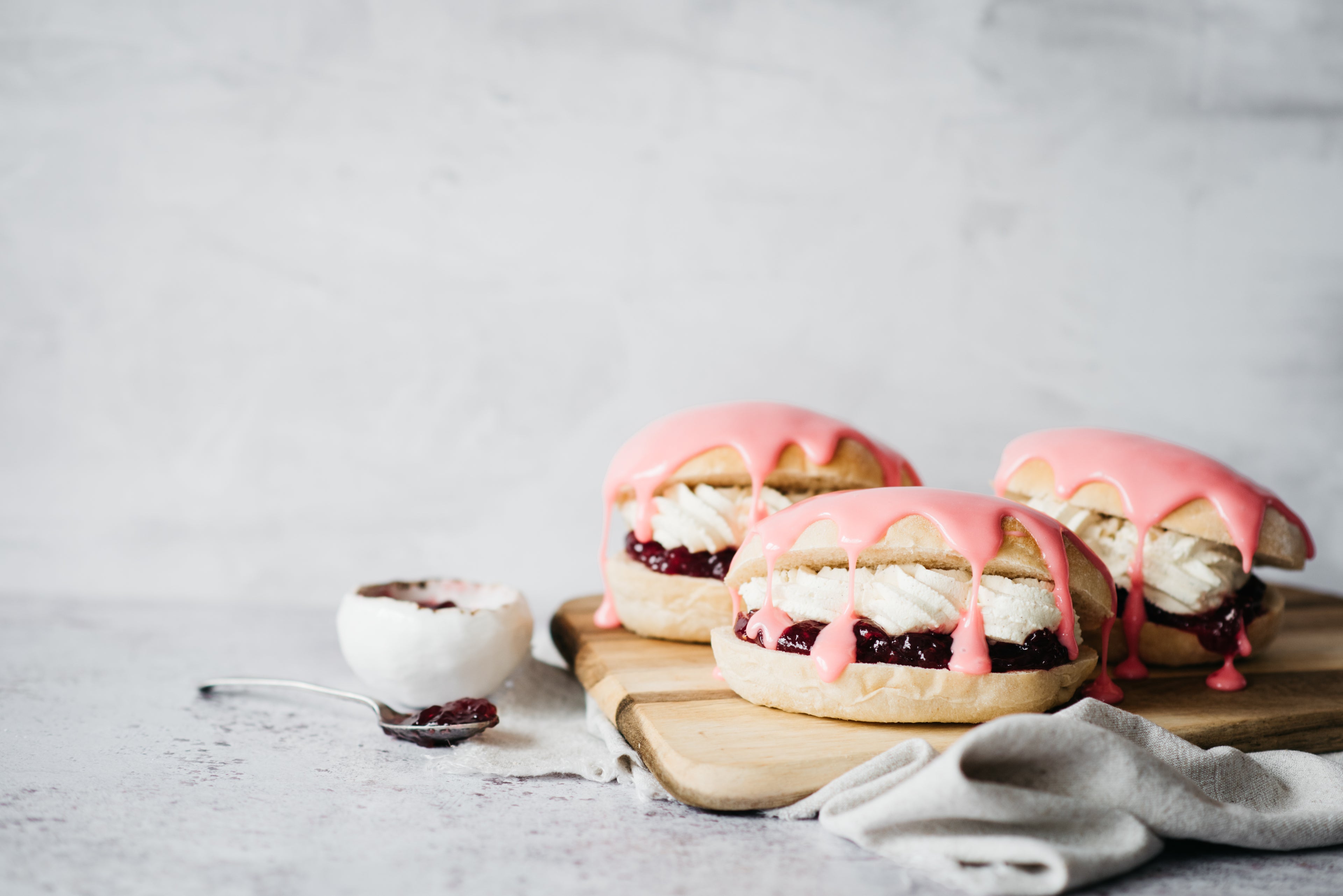 Three Iced Finger Buns on a wooden serving board, drizzled in pink icing next to a bowl of jam and a spoon