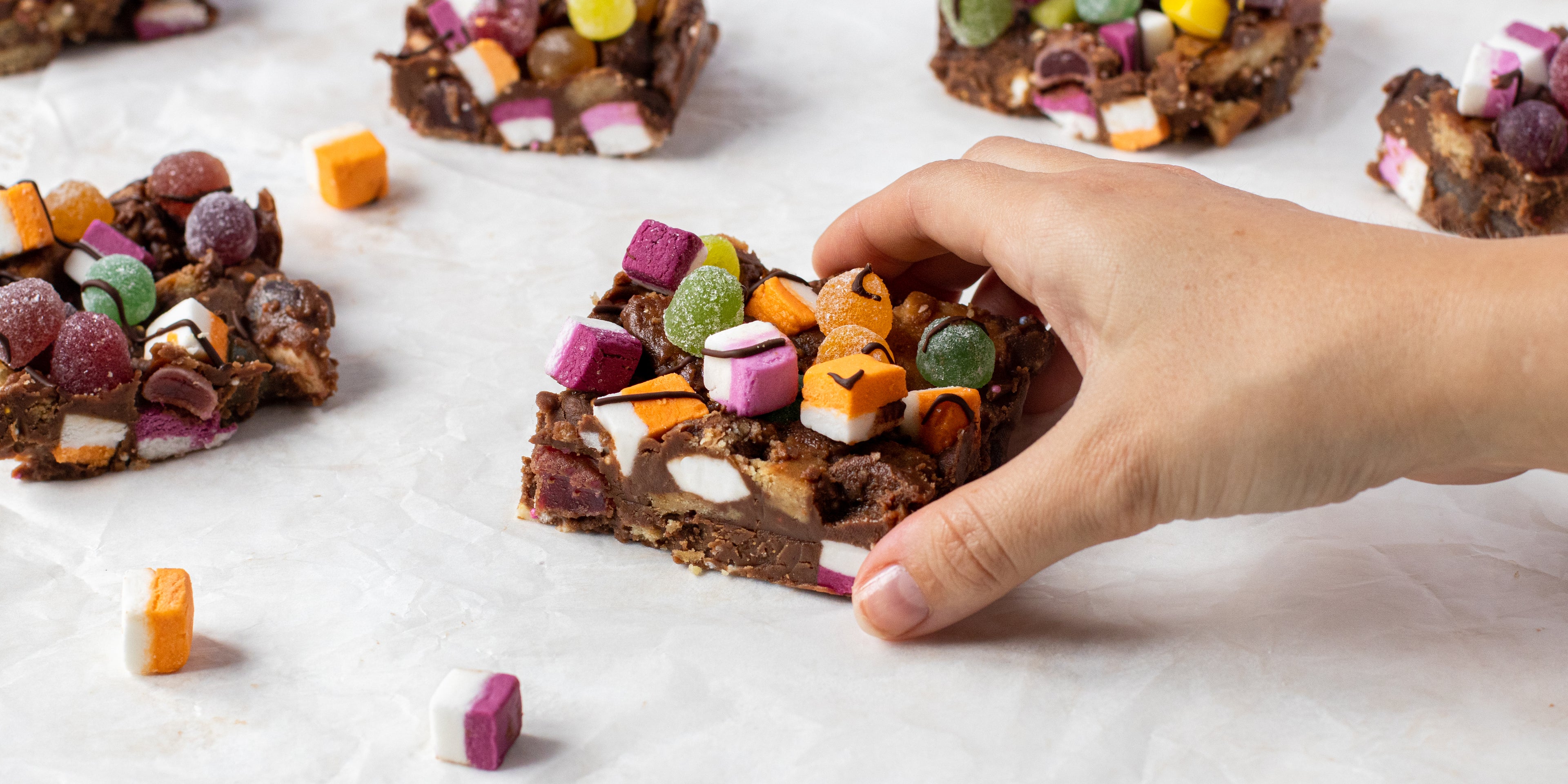 Close up of Sweetie Rocky Road slice, with a hand reaching for it, topped with dolly mixtures
