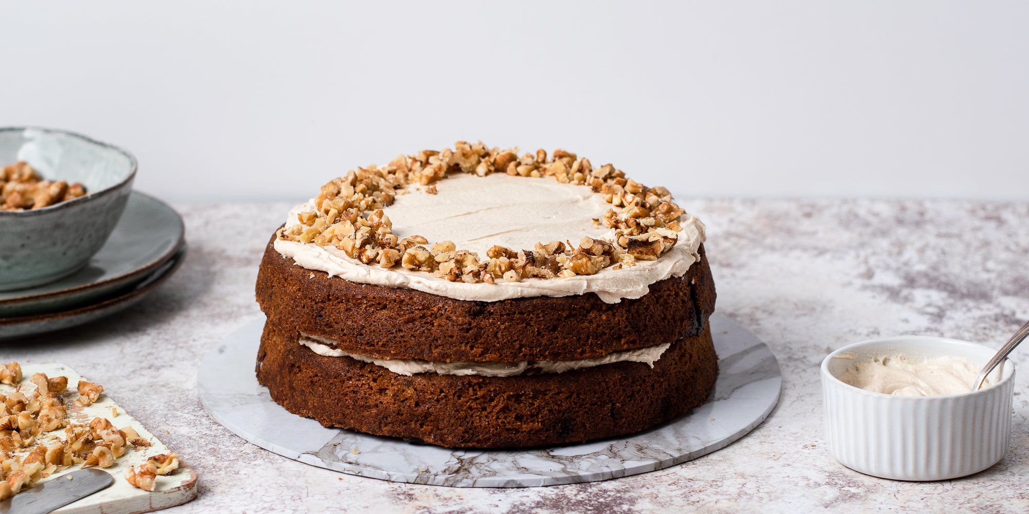 Gluten Free Vegan Coffee Cake on a cake board, next to a ramekin of buttercream and chopping board with chopped nuts