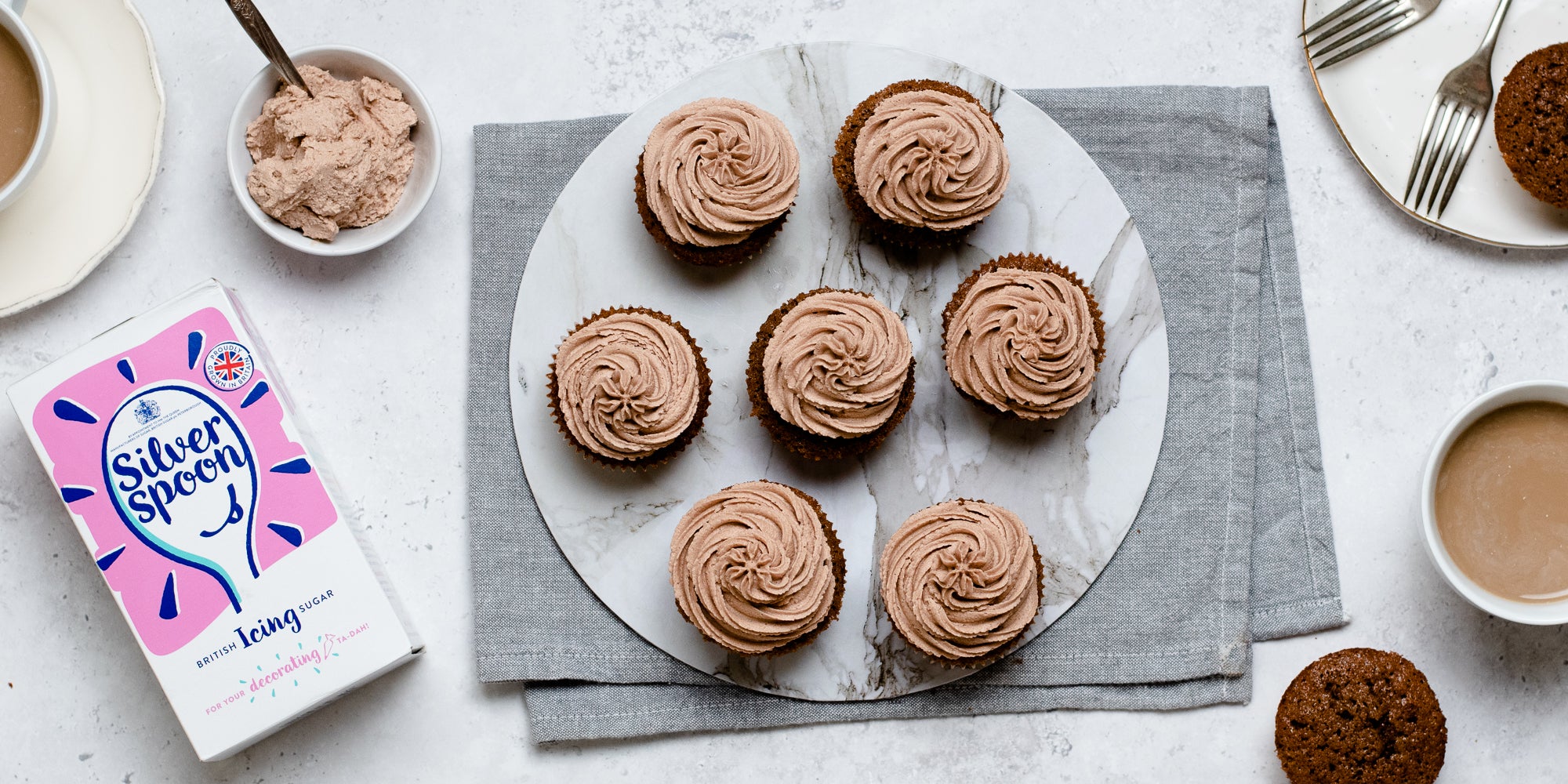 Top view of cupcakes swirled with Dairy Free Vegan Chocolate Buttercream, next to a pack of Silver Spoon Icing Sugar on a linen cloth