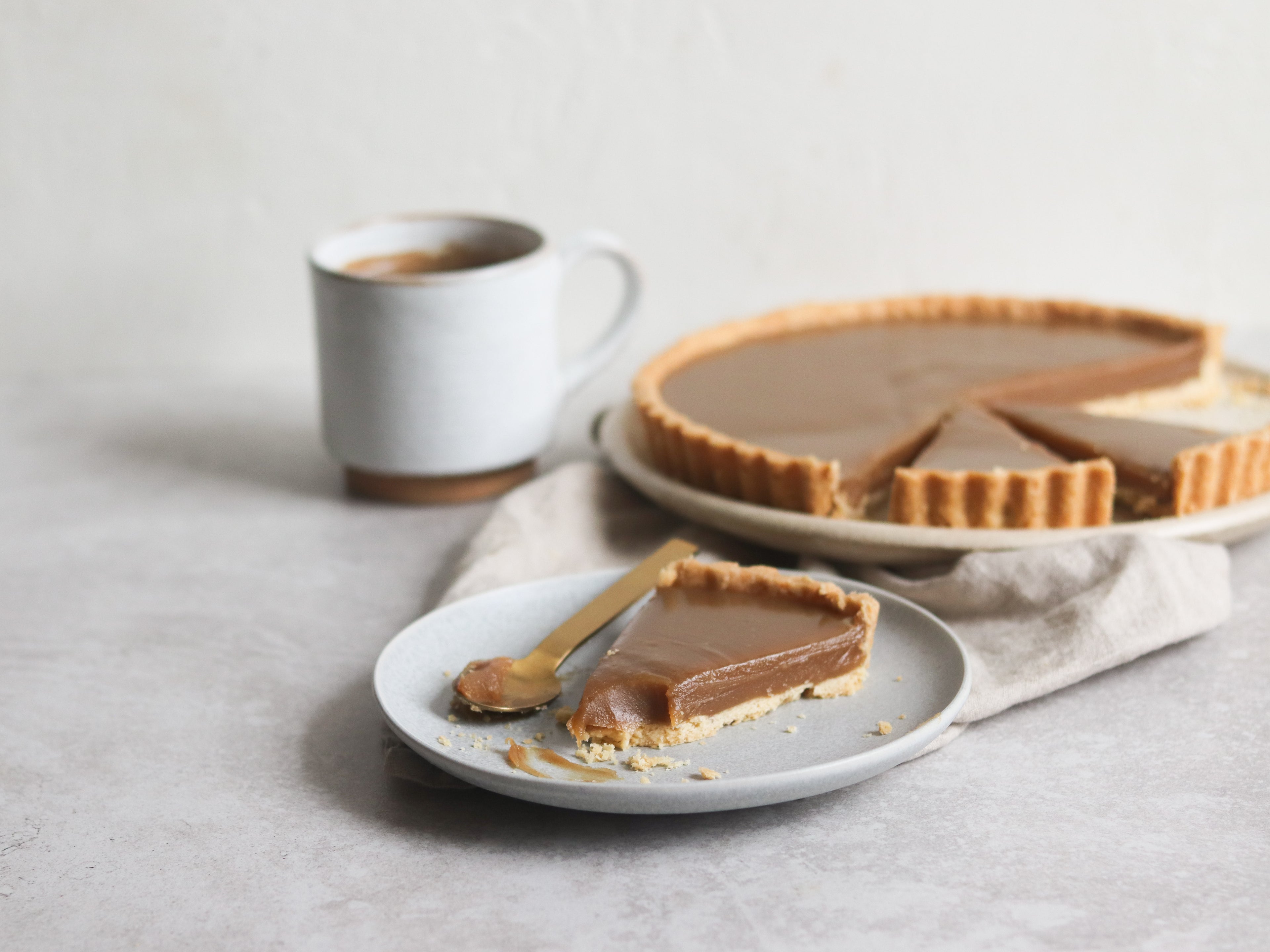 Close up of a slice of Butterscotch Tart, with a piece taken out on a spoon. Cup of tea in the foreground.