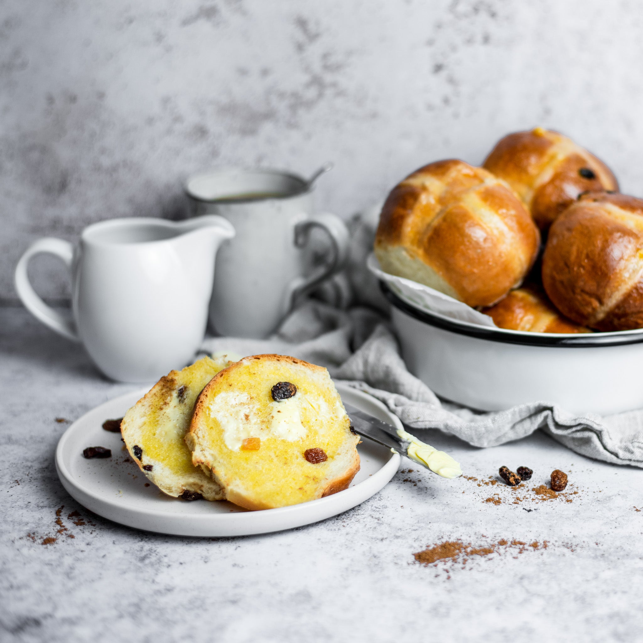 Buttered hot cross buns on a white plate next to a milk jug