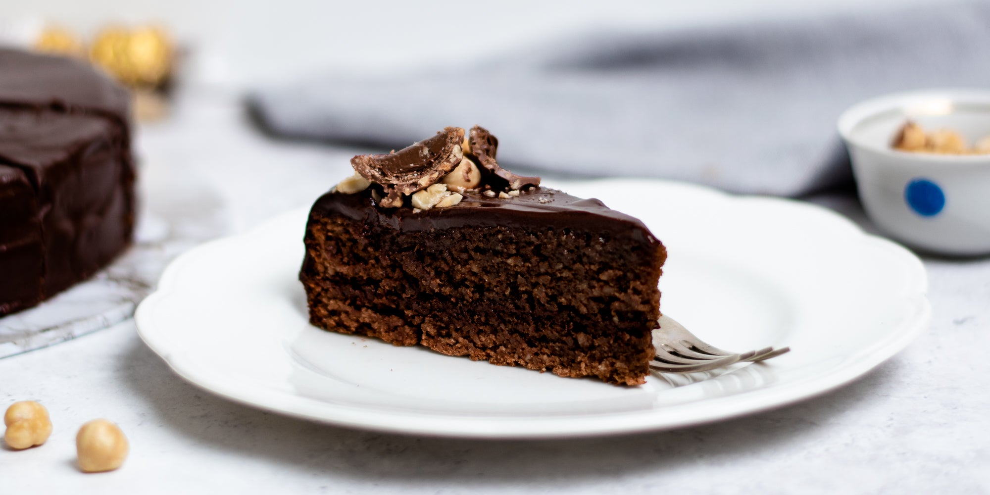 Close up of a slice of Gluten Free Chocolate Truffle Sachertorte served on a plate, scattered in hazelnuts next to a fork