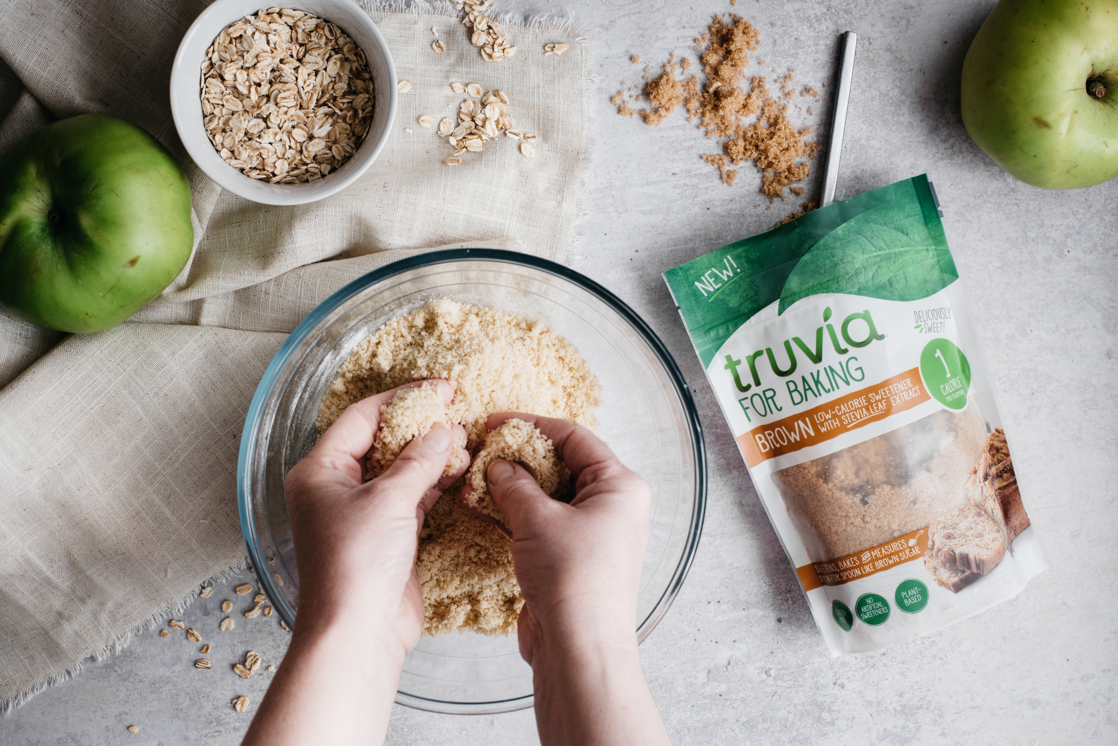 Hands crumbling Mini Apple & Cinnamon Crumble into a bowl, next to a bag of Truvia for Baking Brown.