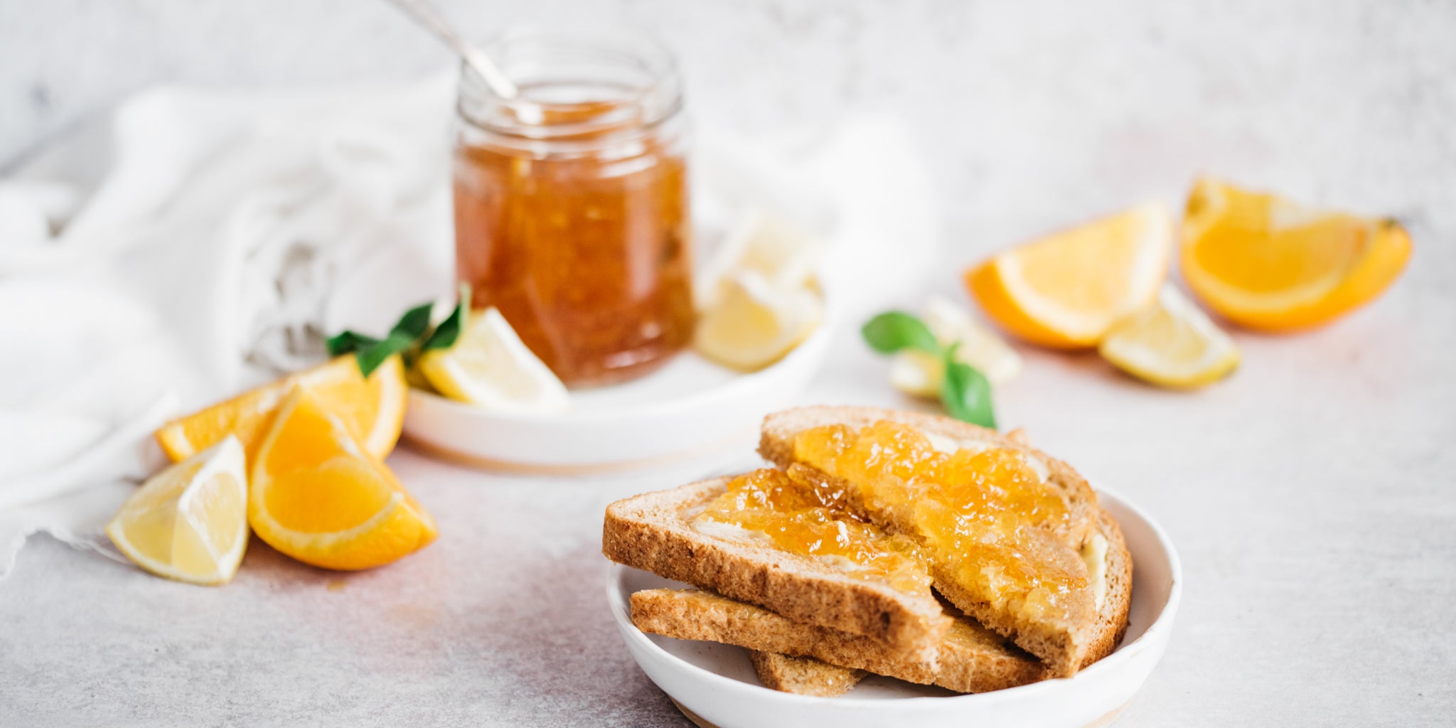 Toast on a white plate spread with marmalade and an open jar of marmalade in background with orange slices