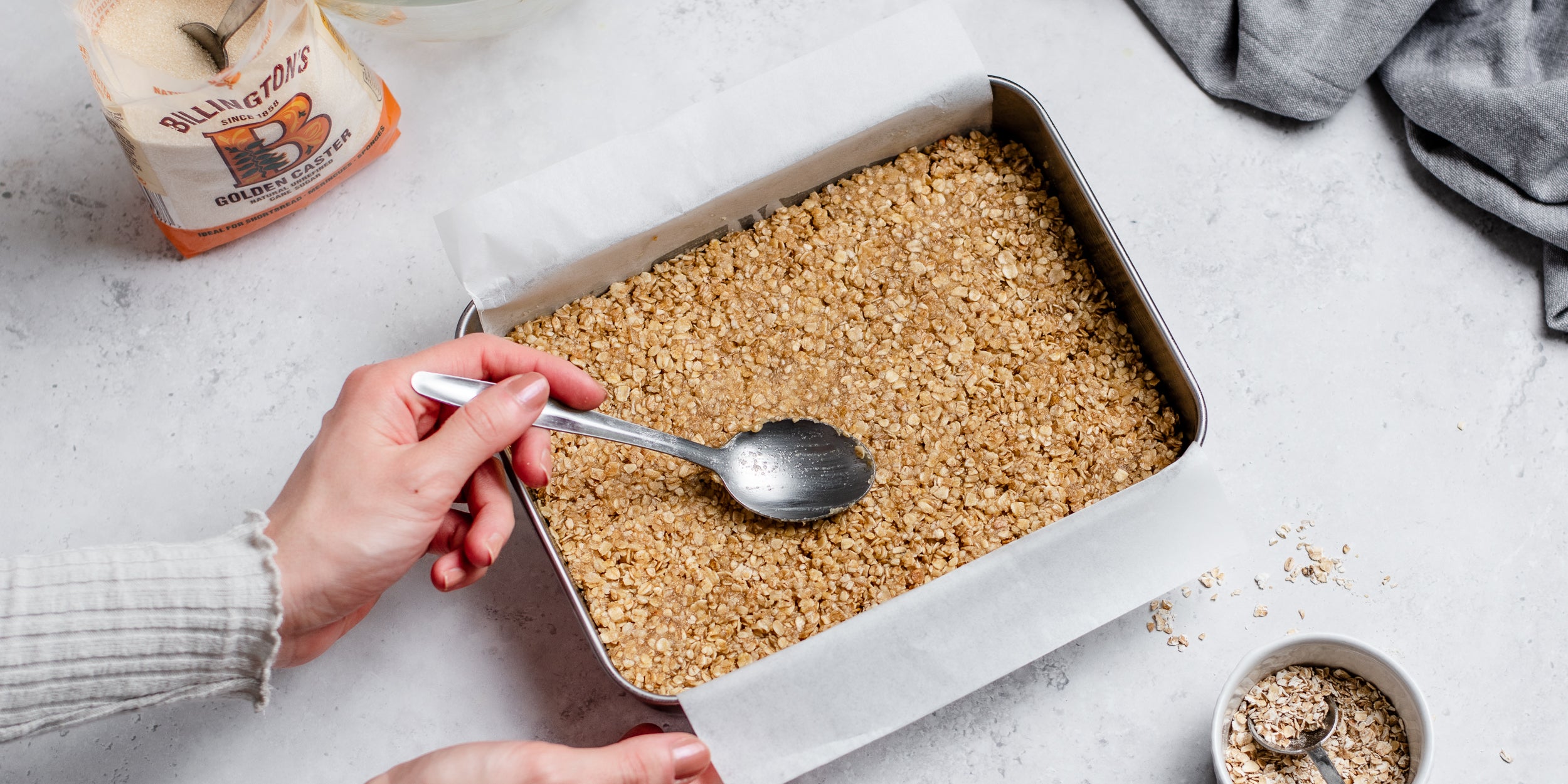 Easy Flapjack being packed into a lined baking tray with a hand holding a spoon. Next to a bag of Billington's Golden Caster Sugar