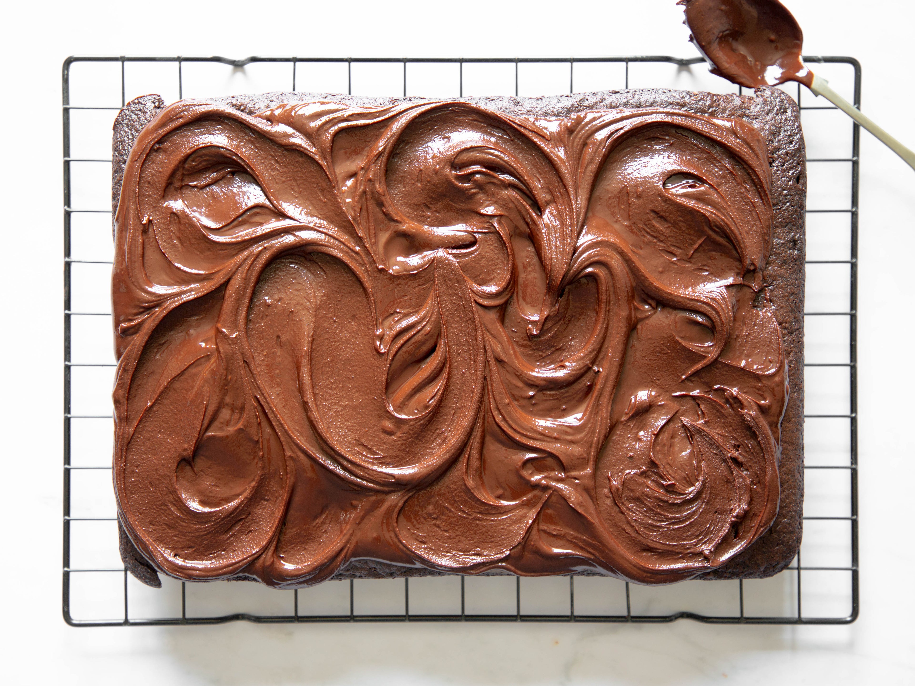 Chocolate traybake covered in chocolate icing on top of a wire cooling rack with a spoon covered in chocolate in top corner