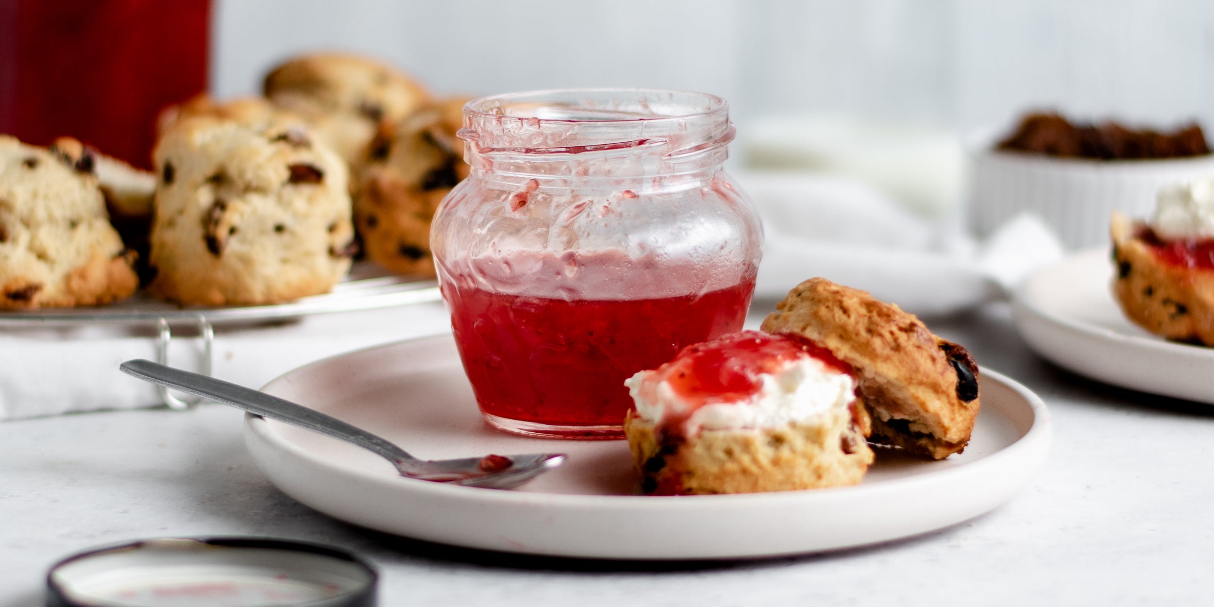Close up of Strawberry Jam next to a scone sliced in half smothered with cream and jam